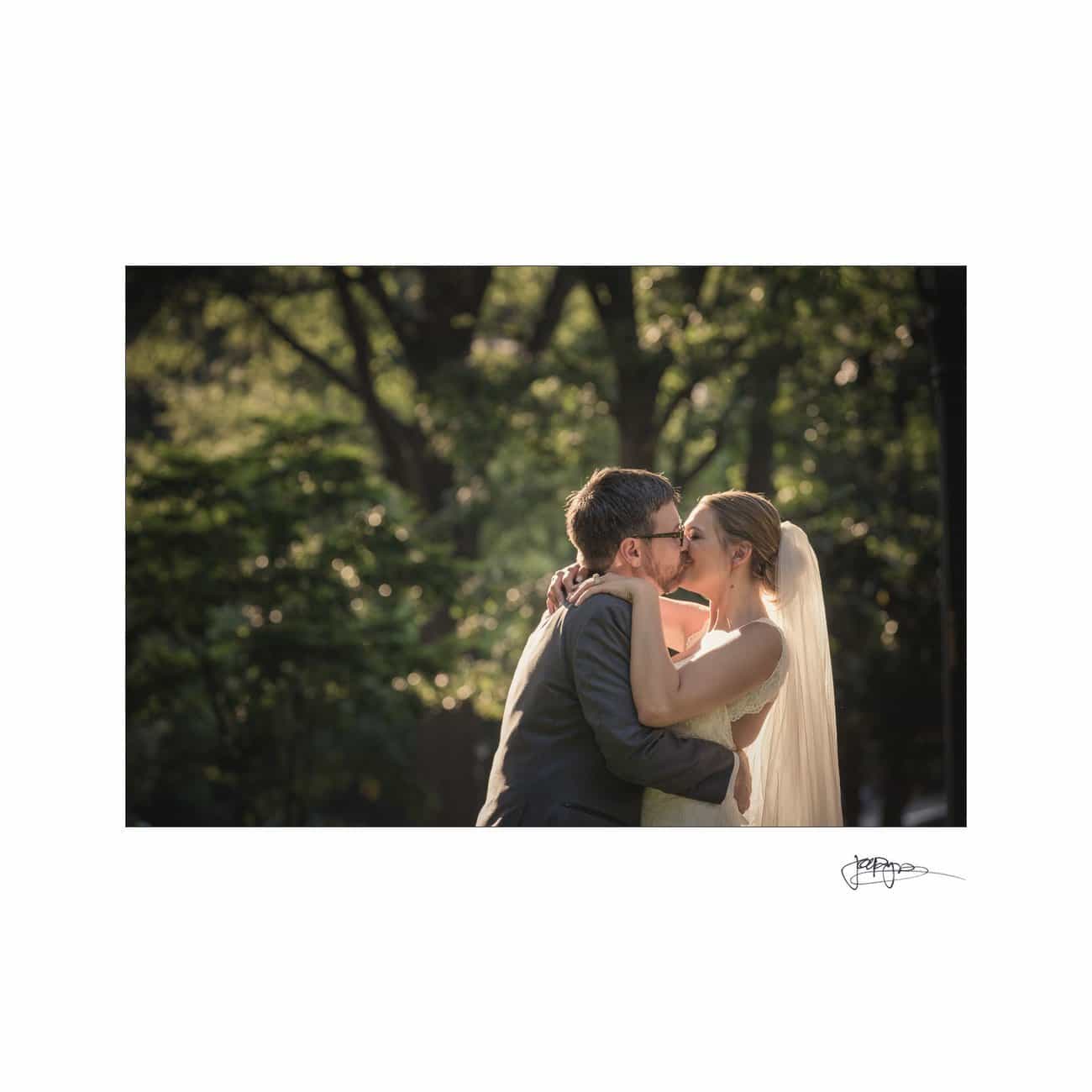 A bride and groom share a kiss outdoors. They are embracing, with the bride wearing a veil and the groom in a suit. Sunlit trees create a soft, natural background.