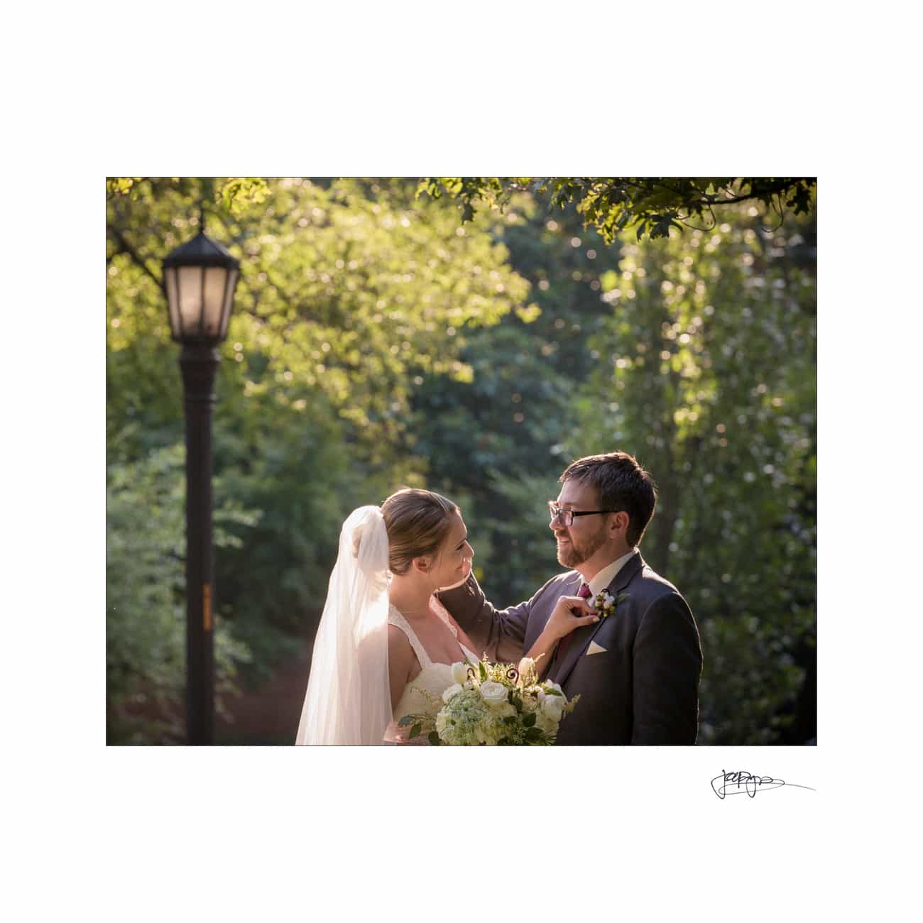 A bride and groom stand together outdoors, surrounded by lush greenery. The bride, in a veil and holding a bouquet, gazes at the groom as she adjusts his boutonniere. A lamppost is visible in the background, and sunlight filters through the trees.