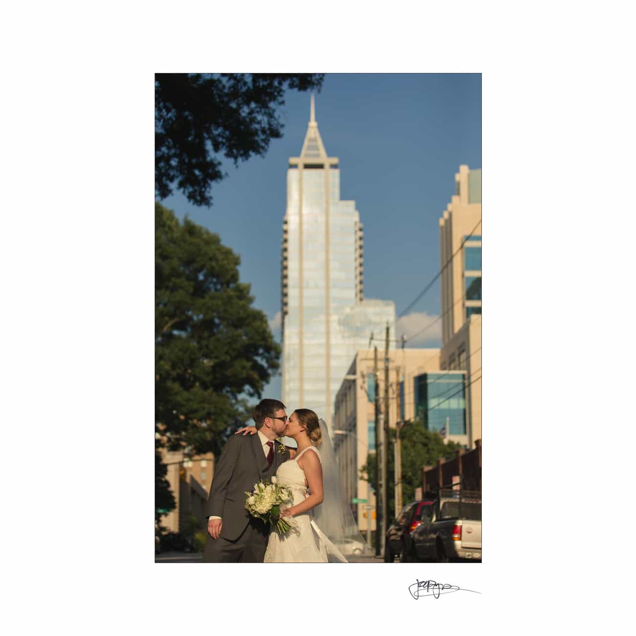 A bride and groom share a kiss on a city street, with a tall building in the background. The bride holds a bouquet of flowers, and the scene is bathed in warm, late-afternoon sunlight. Cars are parked along the street.