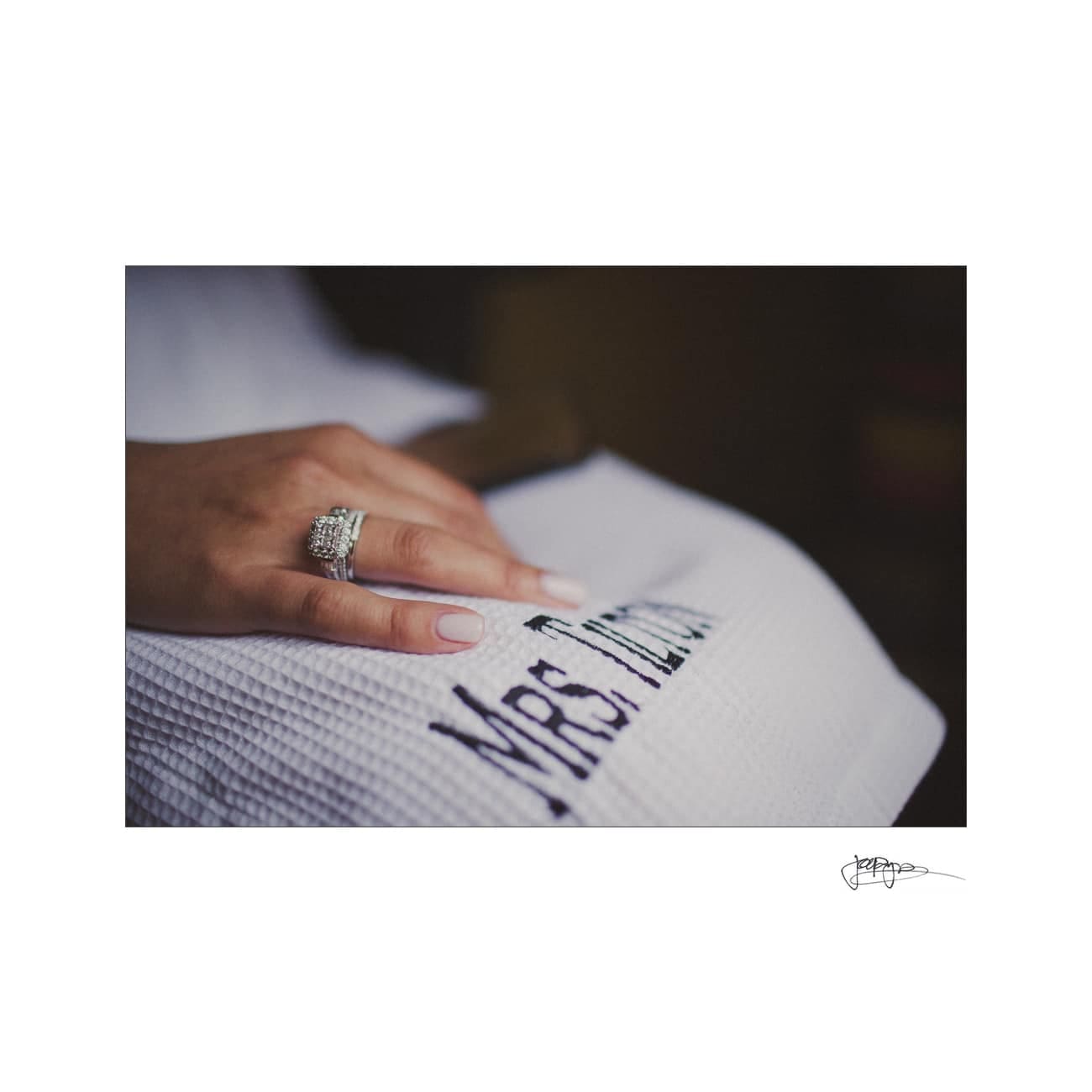 A persons hand with a wedding ring rests on a textured white fabric embroidered with Mrs. The soft lighting and blurred background create a warm, intimate atmosphere.