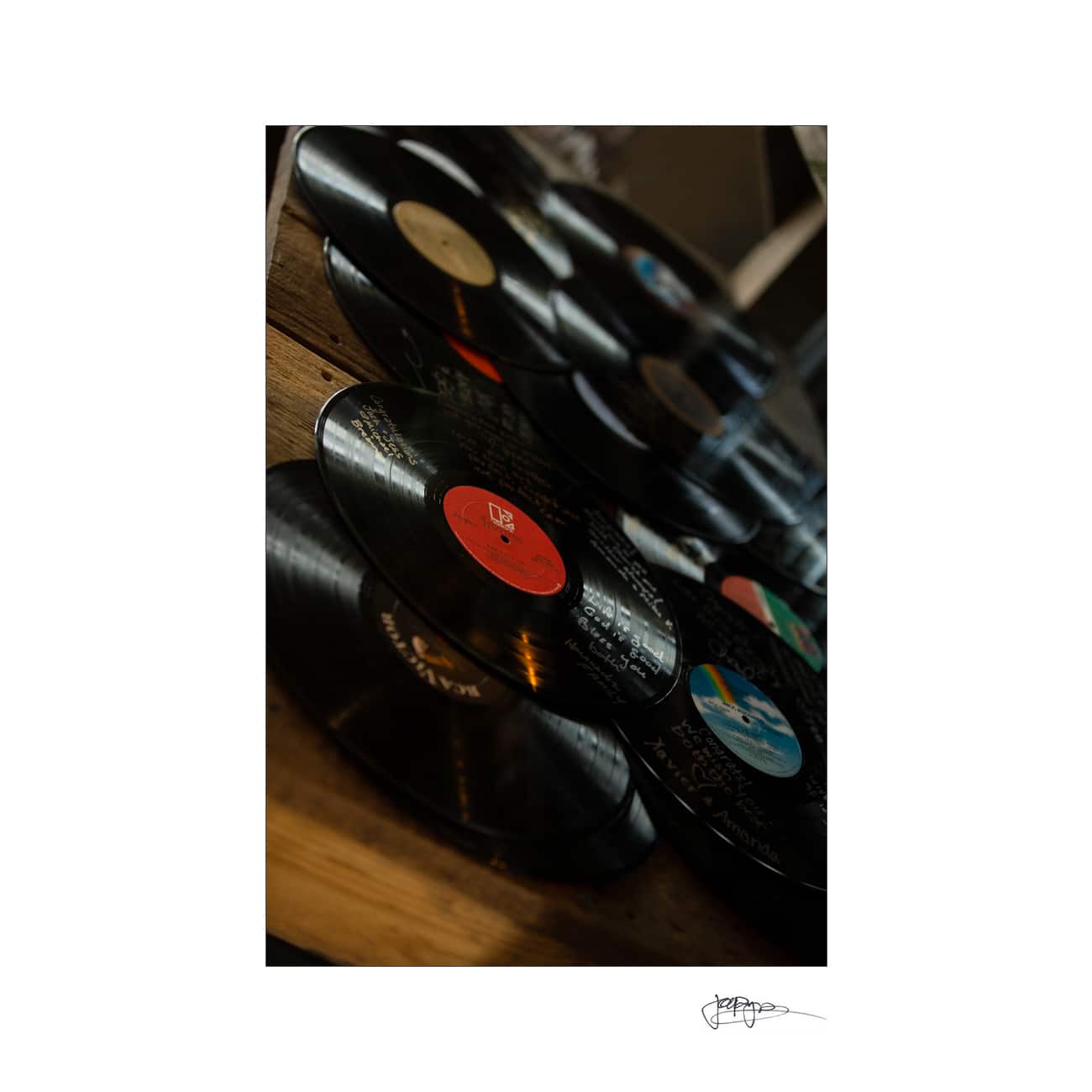 A collection of vinyl records spread out on a wooden surface, featuring various labels and album designs. The angle creates a layered, artistic display with a moody lighting effect.