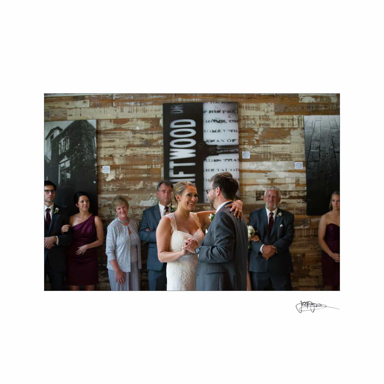 A bride and groom share their first dance in a rustic venue with wooden walls and large black-and-white photos. Bridesmaids, groomsmen, and guests stand around, smiling and watching the couple.