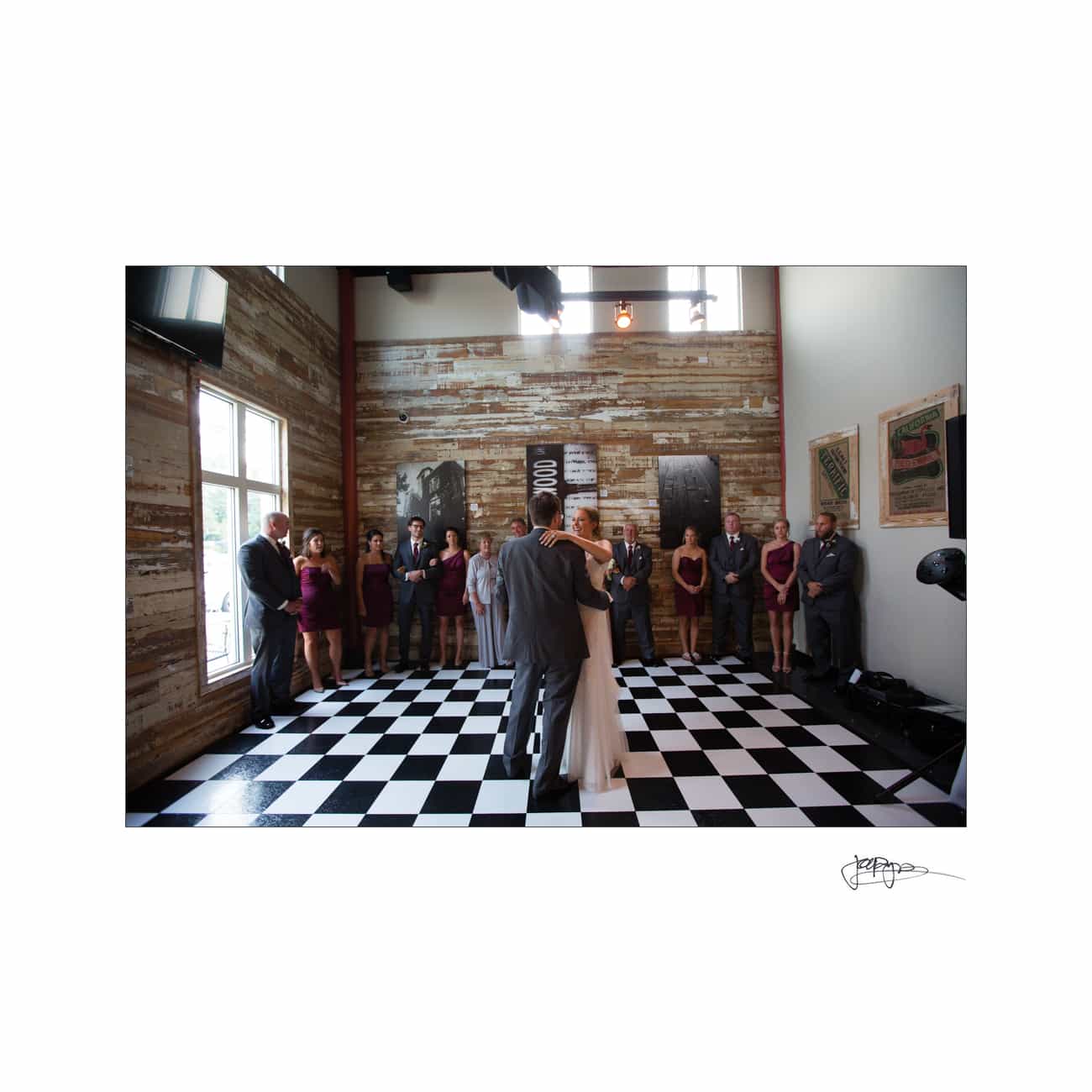 A couple shares their first dance on a black and white checkered floor in a rustic room with wooden walls. Wedding party members stand along the walls, watching.