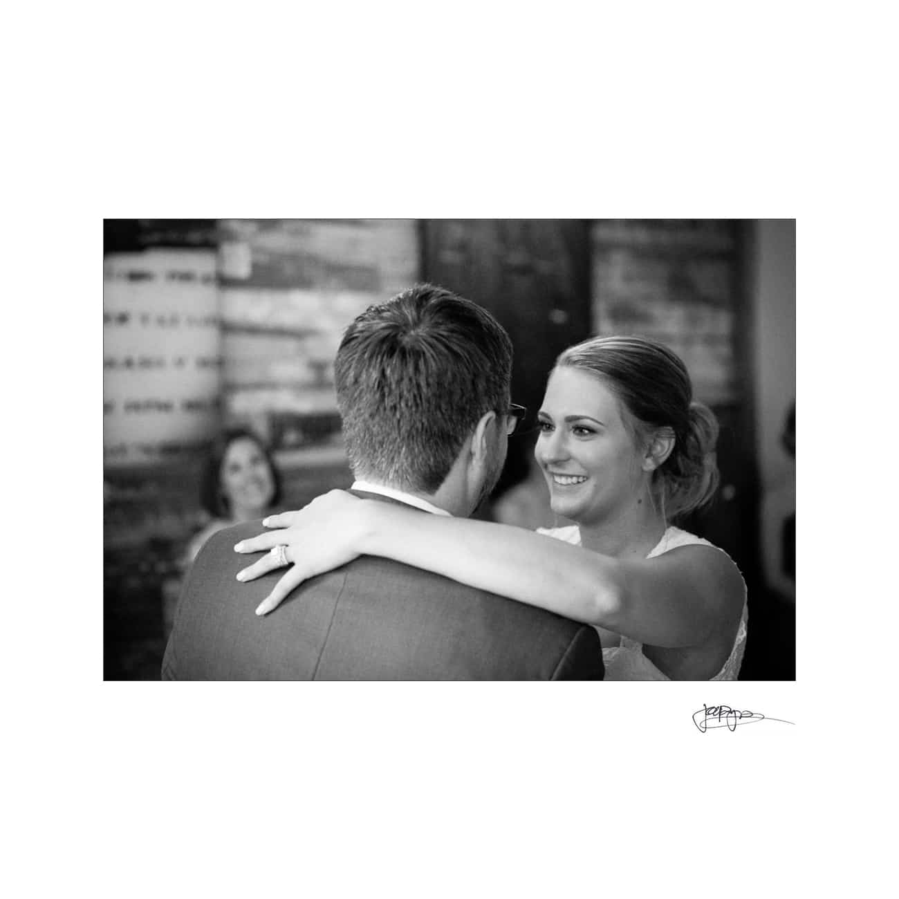 A black and white photo of a couple dancing closely, with the woman smiling as she embraces the man. A person in the background is smiling at them.