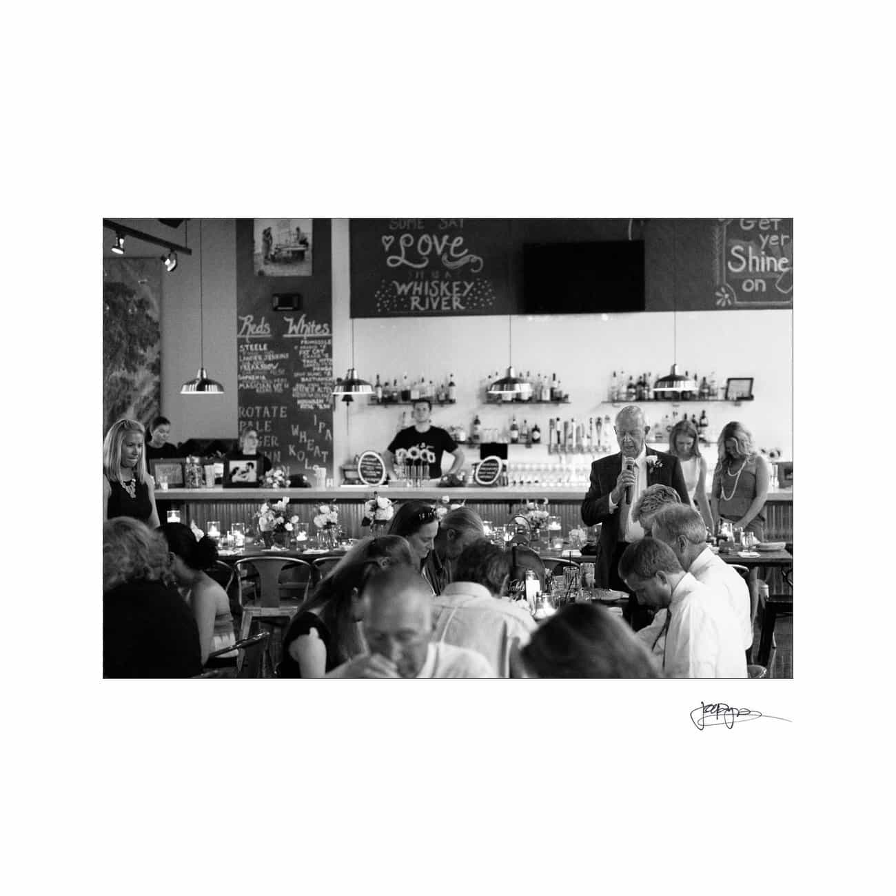 A black and white photo of a lively restaurant scene. A man is speaking to a seated crowd. In the background, two staff members are visible behind the bar. Candles are lit on the tables, creating a warm, inviting atmosphere.