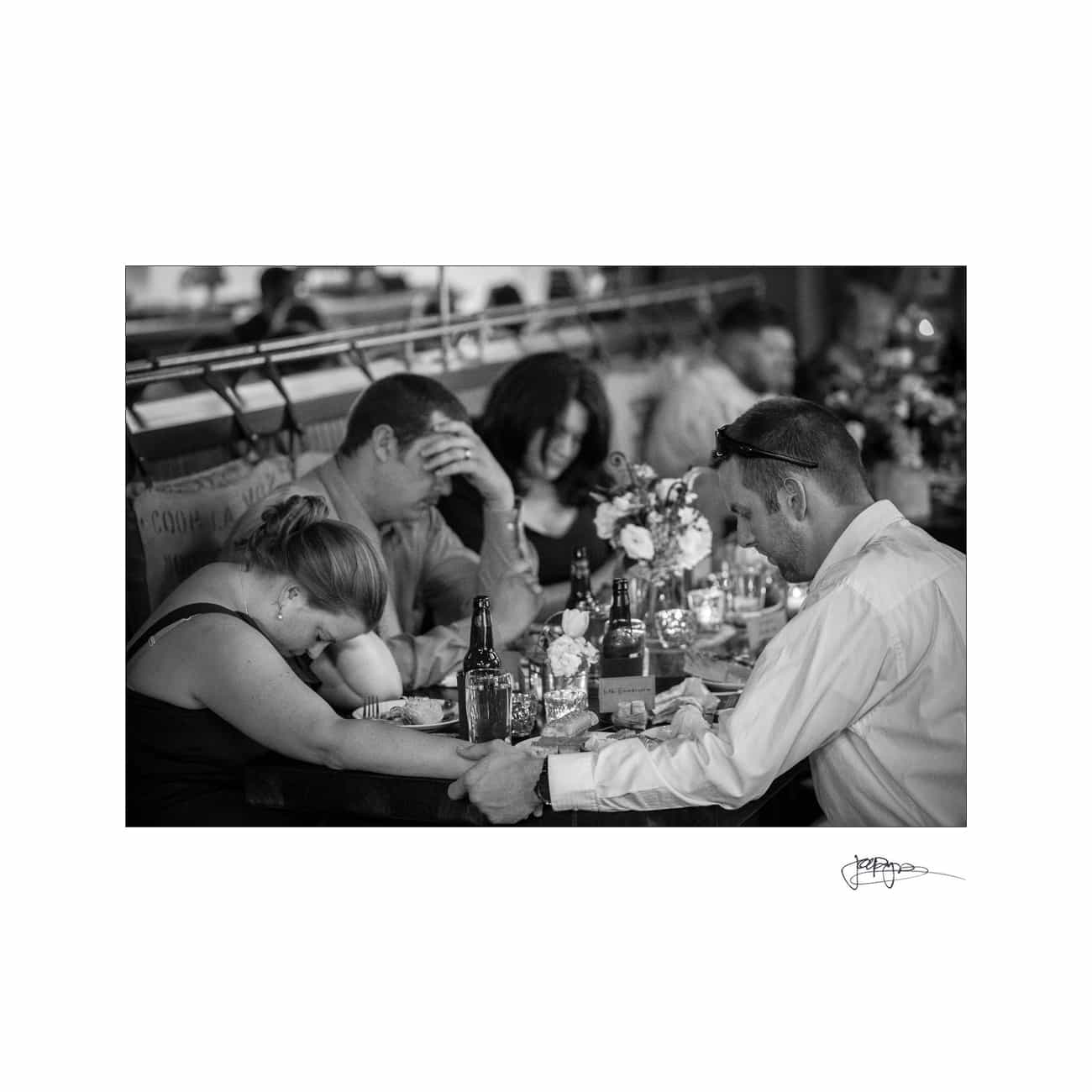 A black-and-white photo of four people sitting at a table in a restaurant. Two people hold hands in the foreground, while the others rest their heads on their arms. The table is decorated with flowers, bottles, and glasses.
