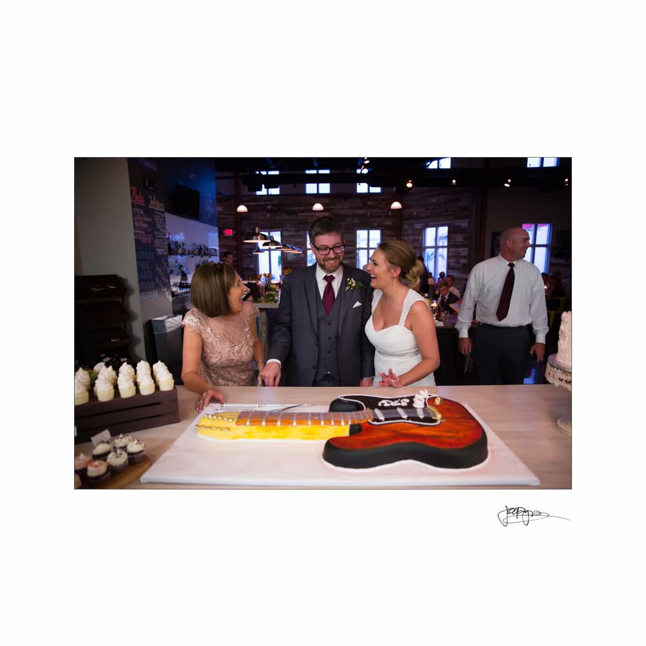 A bride and groom stand with a woman in front of a guitar-shaped cake at their wedding reception. The bride wears a white dress, and the groom is in a suit. The setting is warmly lit with guests and cupcakes in the background.