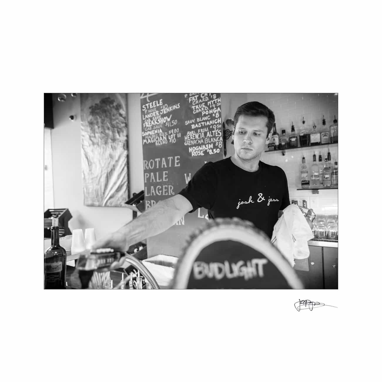 A bartender in a jack & jin shirt serves drinks. The bar has a menu board listing beer types like pale ale and lager. Bottles are displayed on shelves behind him. The image is in black and white.