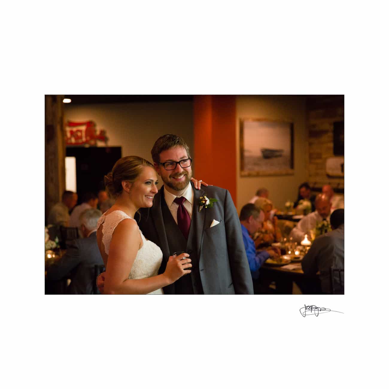 A bride and groom smiling and standing together in a warmly lit room. The bride is wearing a white dress, and the groom is in a suit with a burgundy tie. Guests are seated at tables in the background, engaged in conversation.