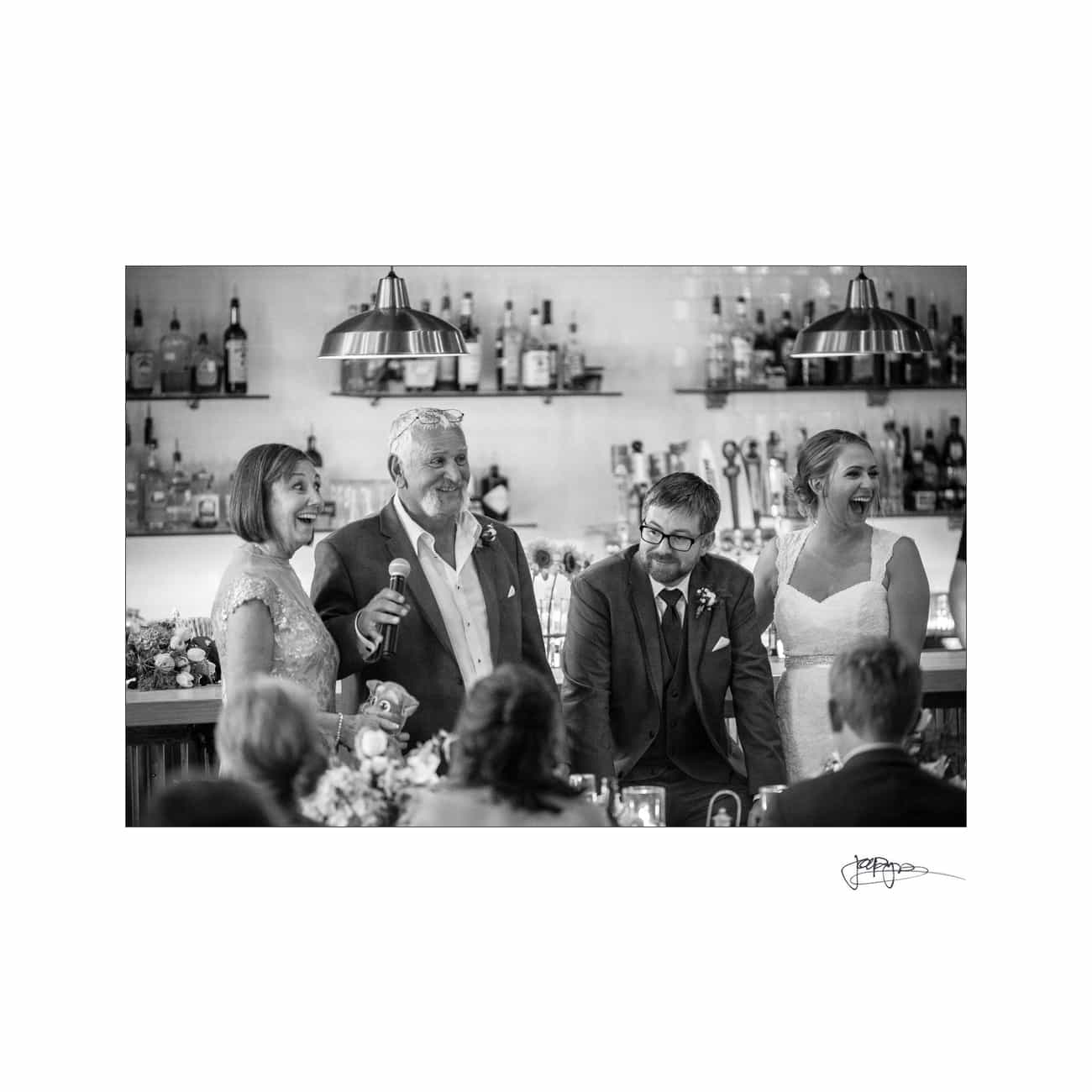 A black and white photo of four people at a wedding reception. Two are standing with microphones, smiling, and dressed in formal attire. The background shows shelves with bottles and hanging lights. Guests are seated in the foreground, partially visible.