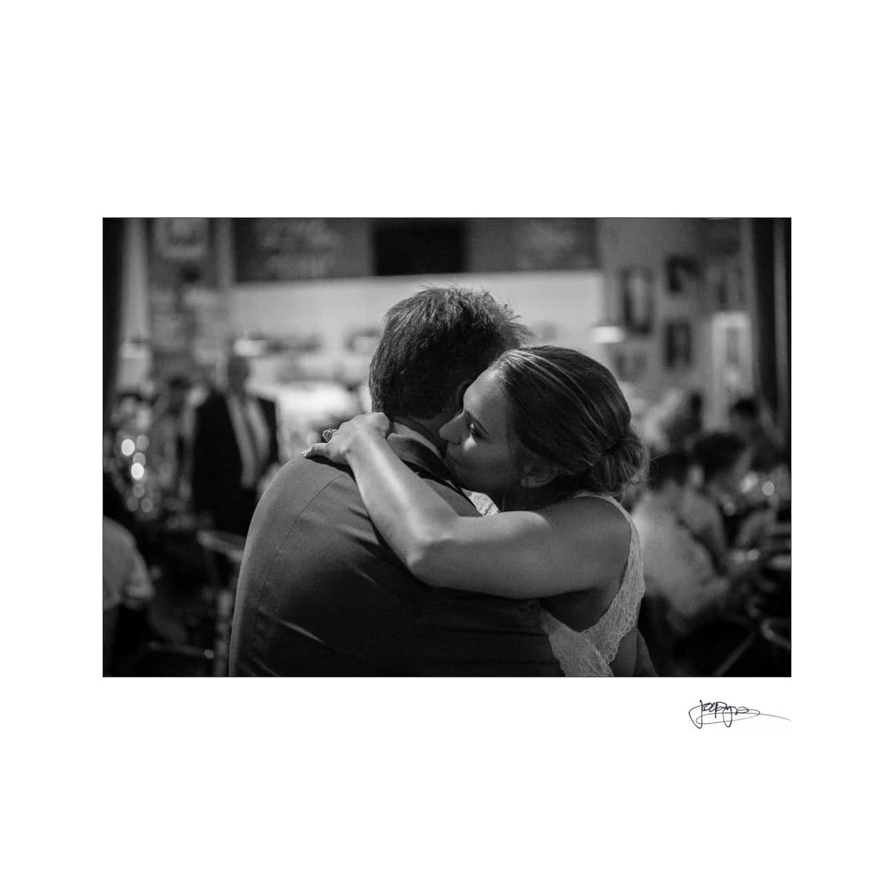 A black and white photo of a couple embracing in a warmly lit room. The woman leans her head on the mans shoulder. The background shows blurred people and decorations, creating an intimate atmosphere.