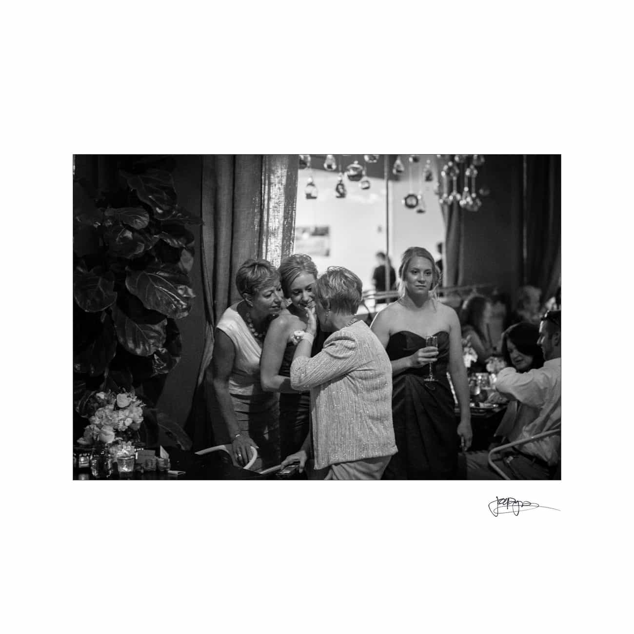A black and white photo of an indoor gathering. Four people are engaged in conversation, with one person holding a drink. The setting is elegant, with plants and decorative lighting in the background.