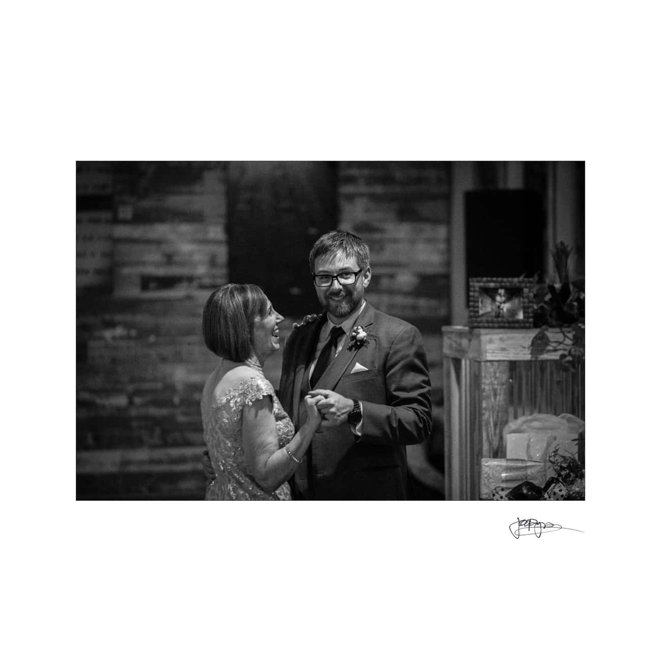 A couple in formal attire dances closely, sharing a joyful moment. They are smiling, with dim lighting and a blurred background that suggests a cozy, intimate setting, possibly at a wedding or special event. The image is in black and white.