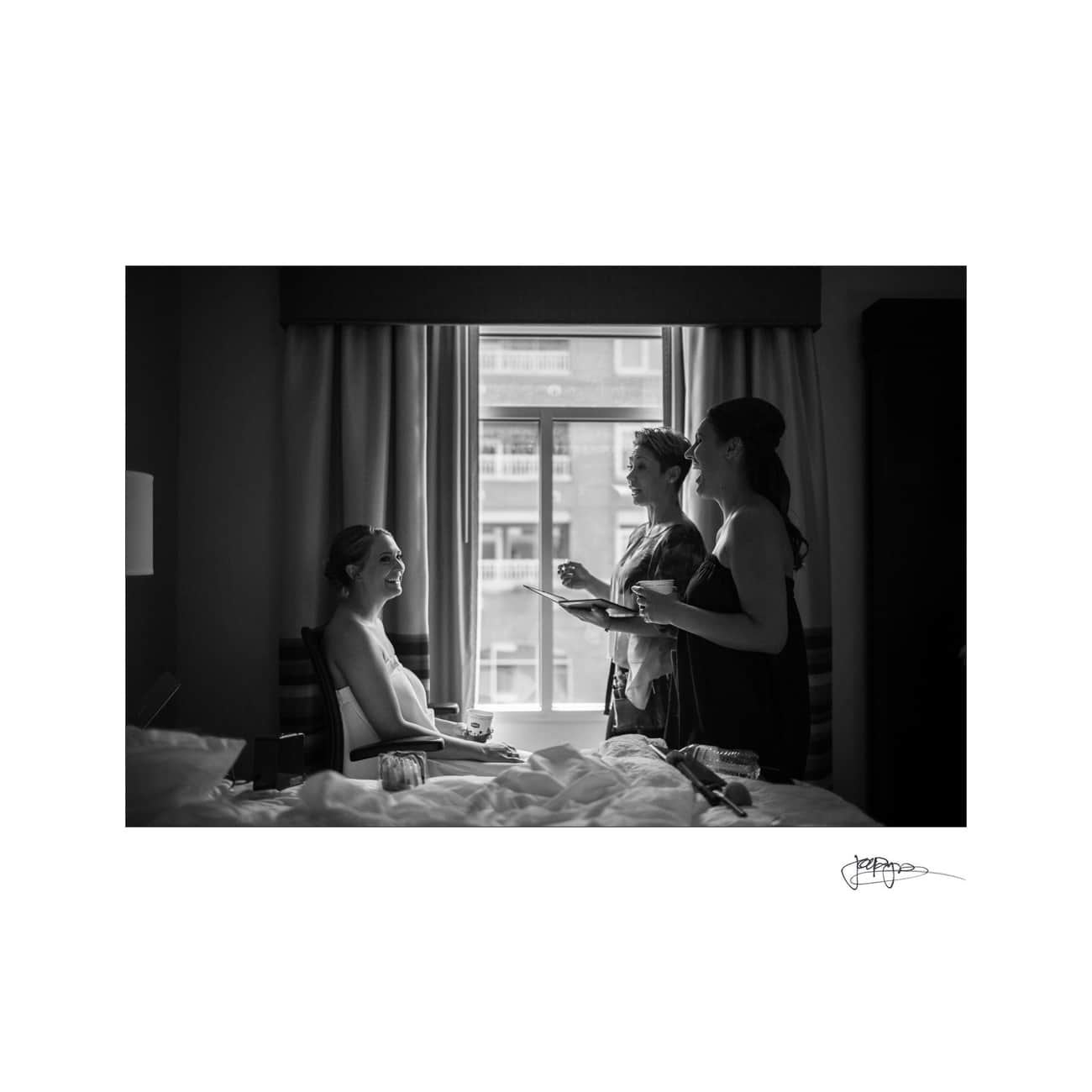 Black and white image of a woman sitting by a window, smiling, as two people assist with her hair and makeup. They stand in a softly lit room with a bed in the foreground and a cityscape visible outside.