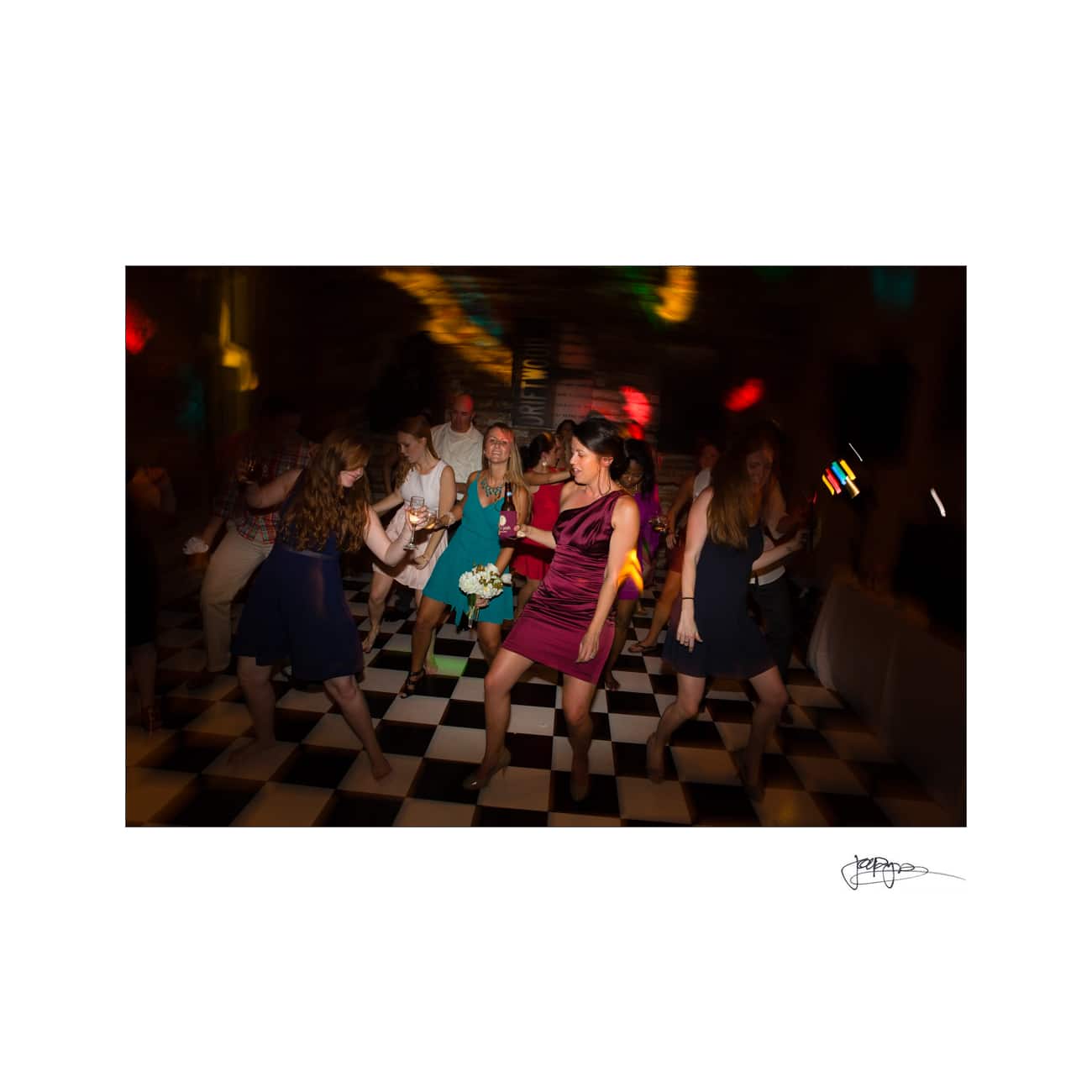 People dancing energetically on a black and white checkered floor, with colorful lights creating a lively atmosphere. A woman in a red dress stands in the foreground holding a drink.