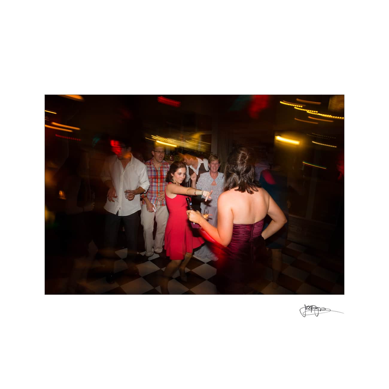 A group of people enjoying a dance party. The scene is lively, with blurred lights and motion capturing the energy. A woman in a red dress is at the center, surrounded by others on a checkered floor.