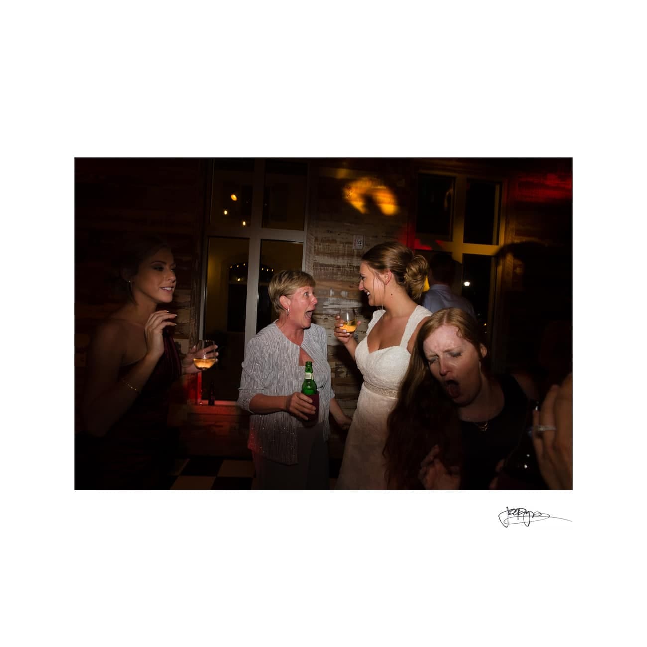 A group of people joyfully celebrating at a party. A woman in a white dress stands in the center holding a drink, surrounded by others with drinks, laughing and engaging in animated conversation in a warmly lit room.
