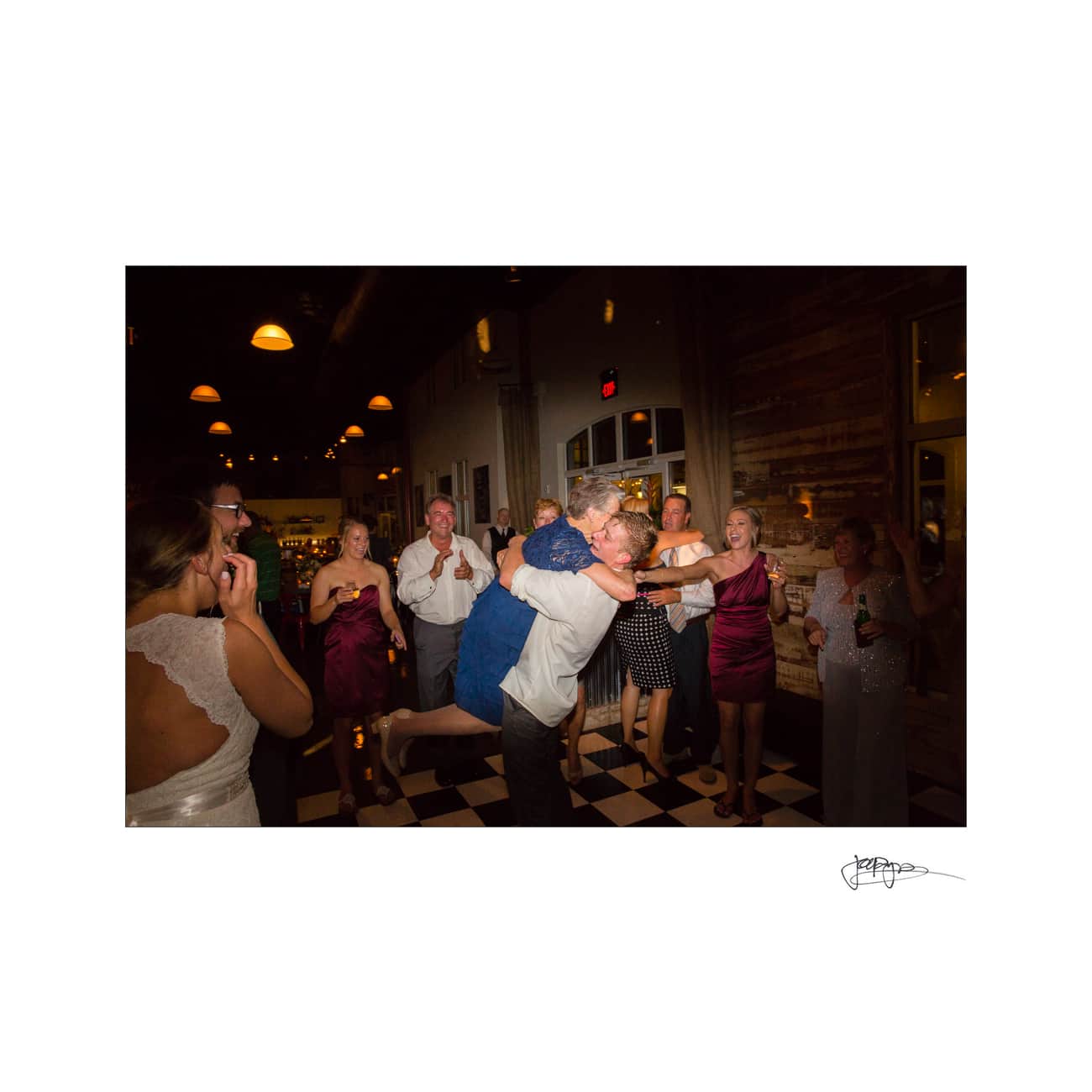 A joyful wedding reception scene with guests dancing on a checkered floor. A couple embraces in the center, surrounded by smiling friends in formal attire, under warm lighting.