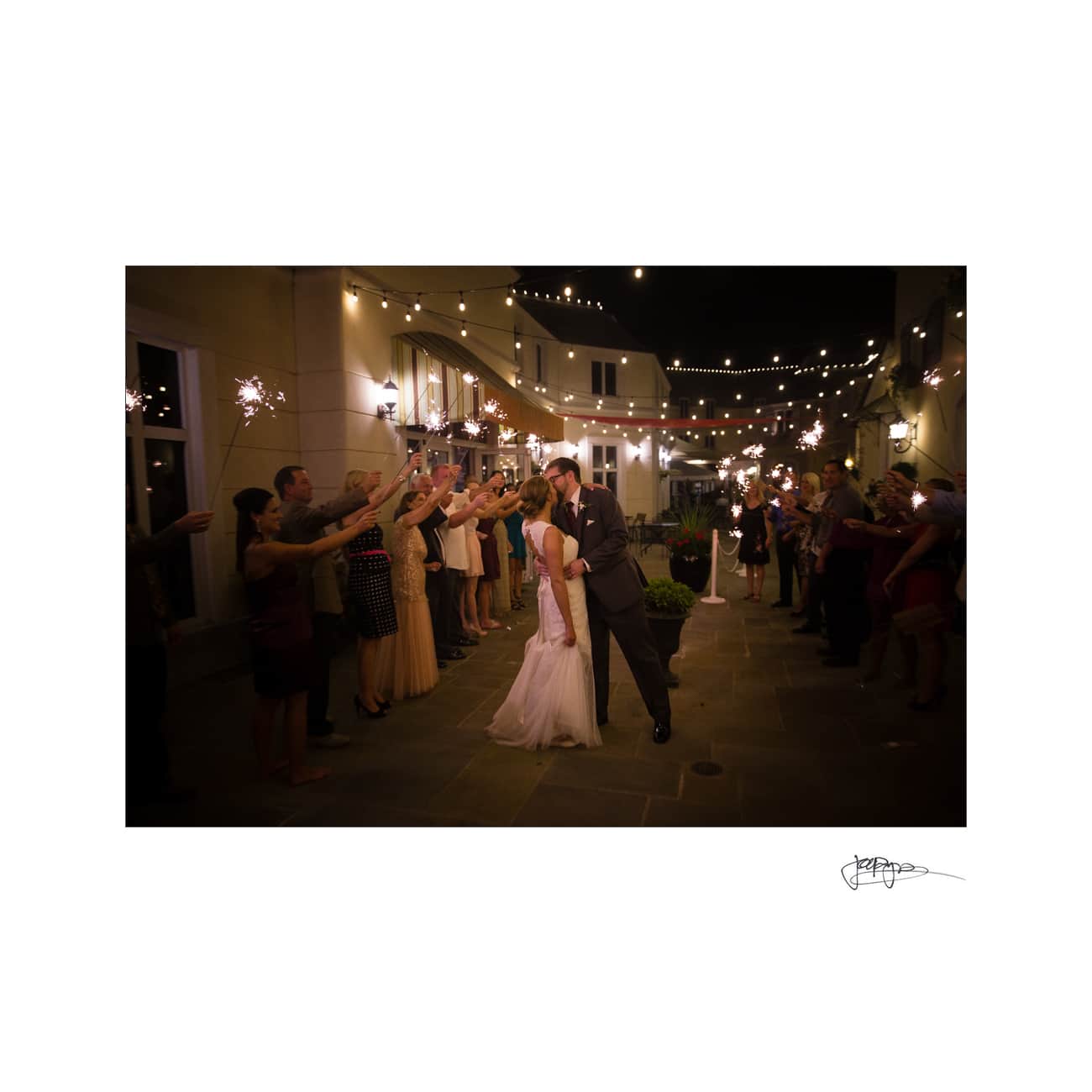 A bride and groom share a kiss at night under string lights. They are surrounded by guests holding sparklers, forming a celebratory arch. The scene is festive and romantic.