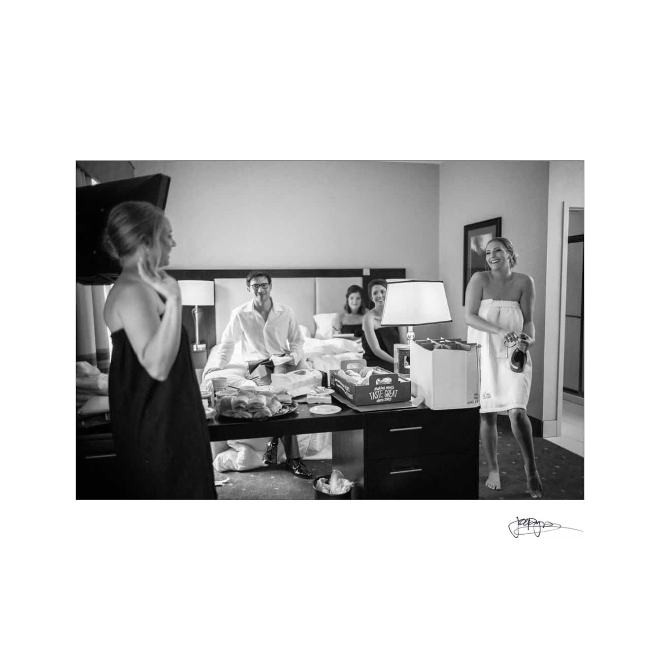 A black and white photo of a hotel room with four people enjoying a relaxed gathering. Two people sit on a bed surrounded by takeout food, while two others stand by, laughing and engaged in conversation. A wine box is visible on the table.