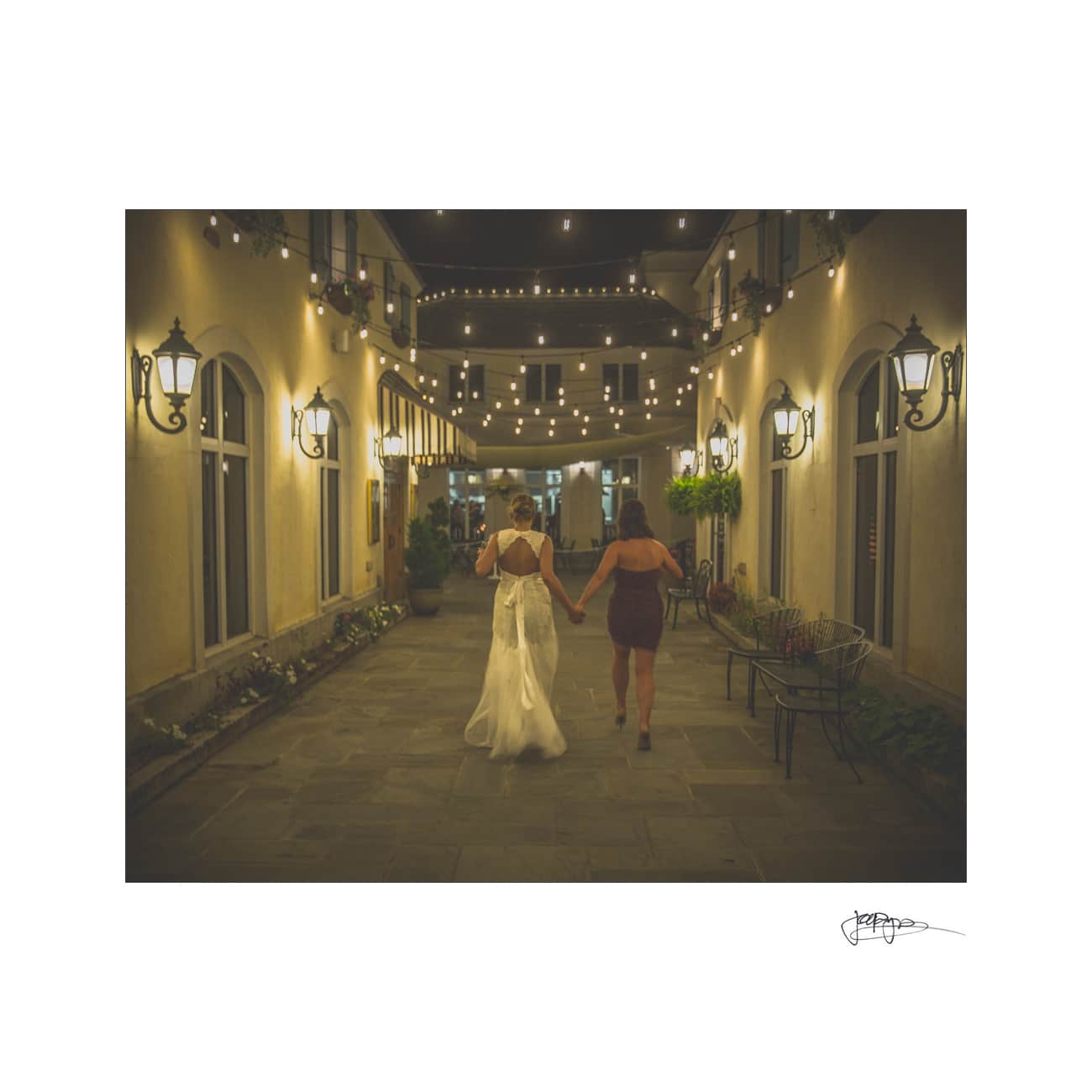 Two people holding hands, walking down a dimly lit, elegant path adorned with string lights and lanterns. One is wearing a white dress, the other a dark outfit. The scene exudes a romantic evening atmosphere.