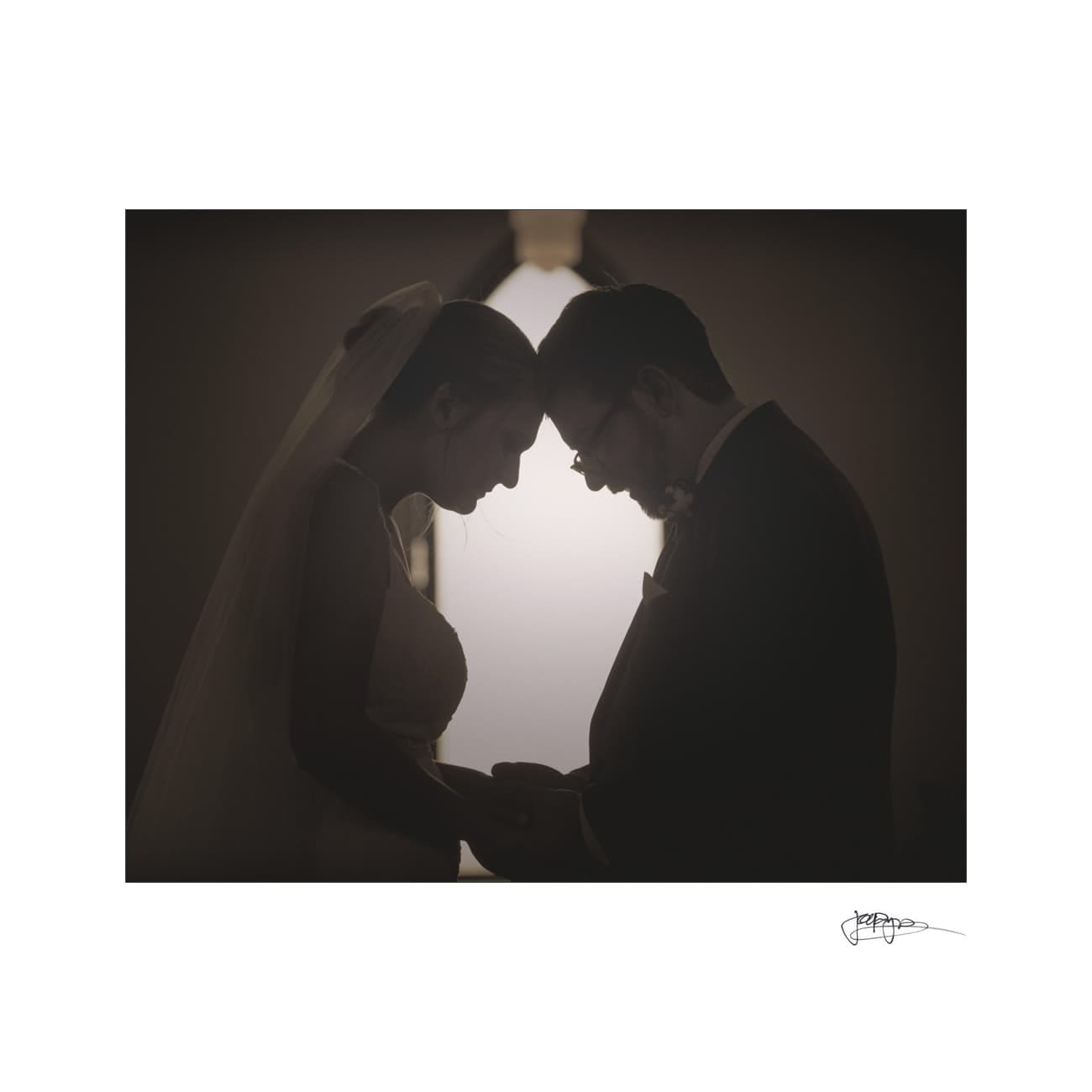 Silhouetted couple in wedding attire stand facing each other, holding hands beneath a soft light. The bride wears a veil, and the groom is in a suit and bow tie, creating a romantic and intimate atmosphere.