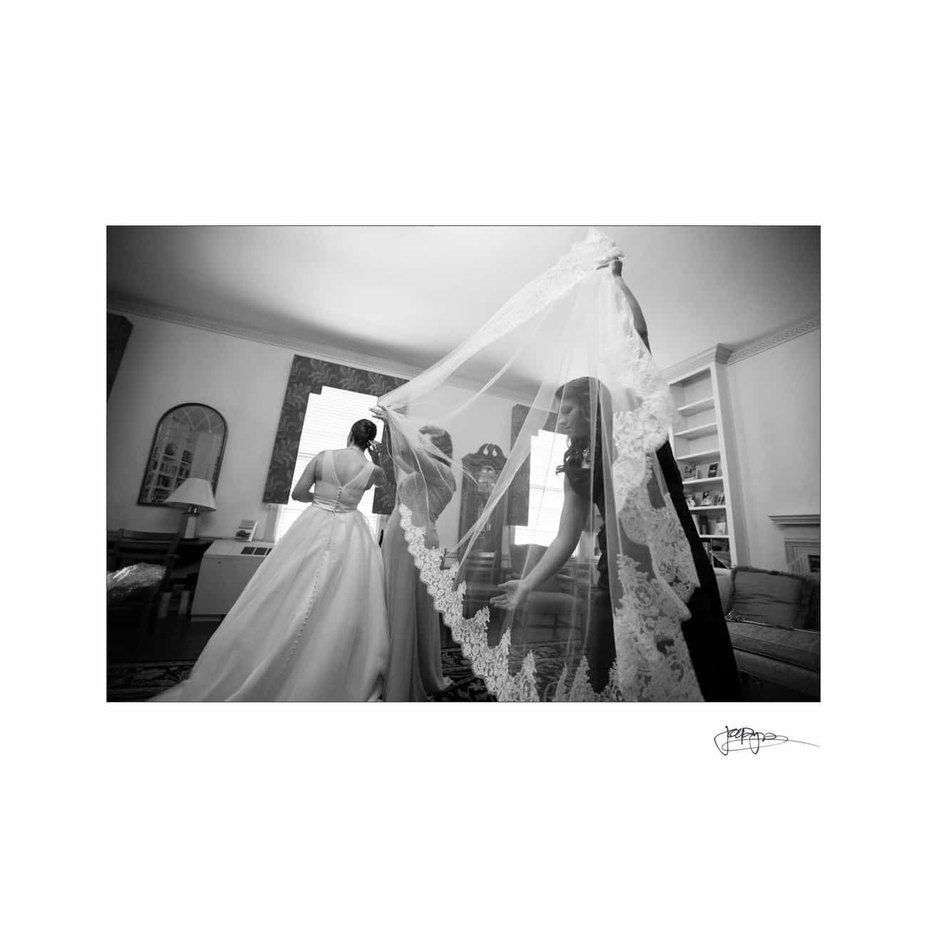 chapel hill wedding photographer - alexandra & john A black and white photo of a bride standing in a living room, wearing a long wedding gown. Two people are helping arrange her veil, which features intricate lace patterns. The room has large windows and bookshelves.