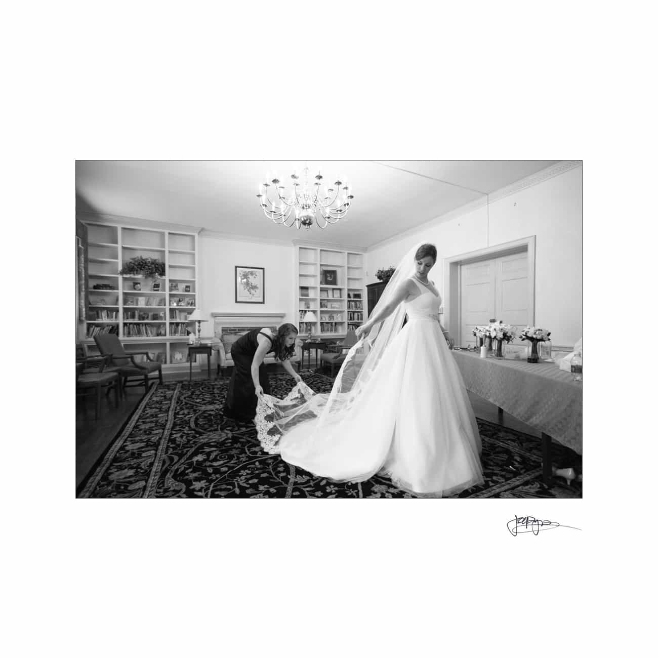 chapel hill wedding photographer - alexandra & john A bride in a wedding gown stands in a room with bookshelves and a chandelier. Another person crouches to adjust the train of her dress. The rooms elegant decor and furnishings add to the scenes formal atmosphere. The image is in black and white.