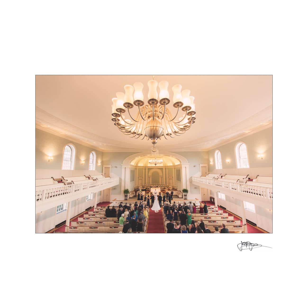 chapel hill wedding photographer - alexandra & john A couple stands at the altar in a large church as guests watch seated in pews. The grand interior features an elaborate chandelier and tall, arched windows that illuminate the scene.