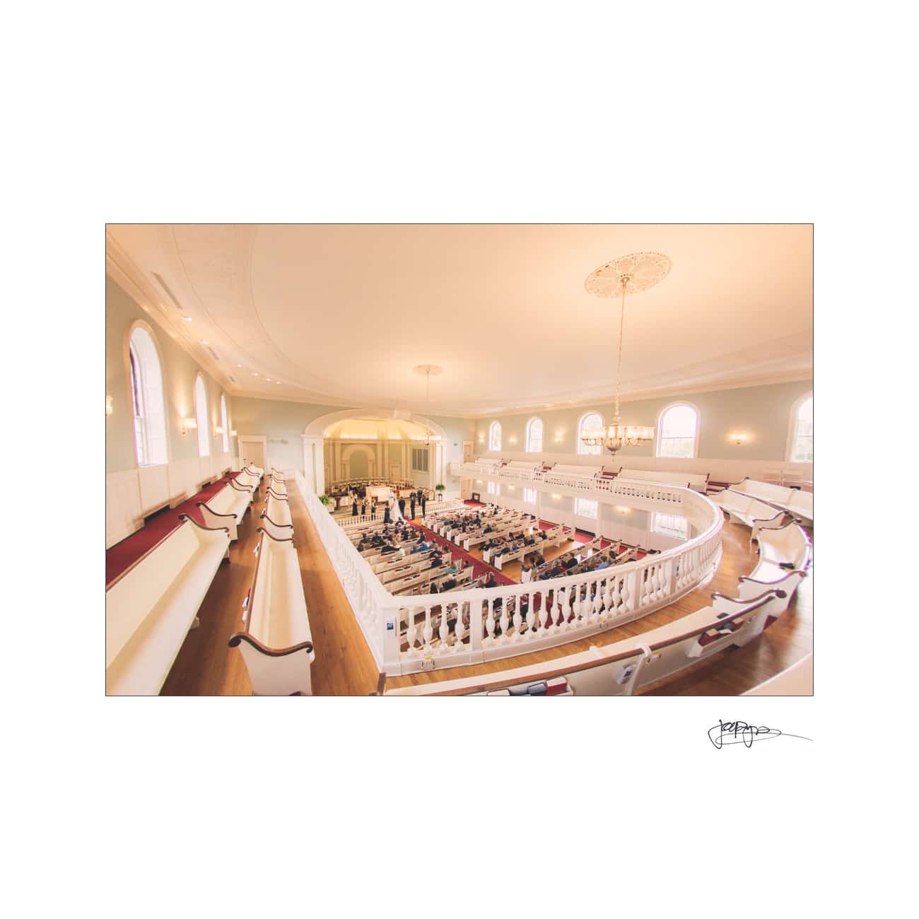 chapel hill wedding photographer - alexandra & john A fisheye-lens view of an elegant church interior with a high ceiling, large chandelier, and arched windows. The congregation is seated in pews, and theres a focus on the central stage area. Light streams in, creating a warm atmosphere.