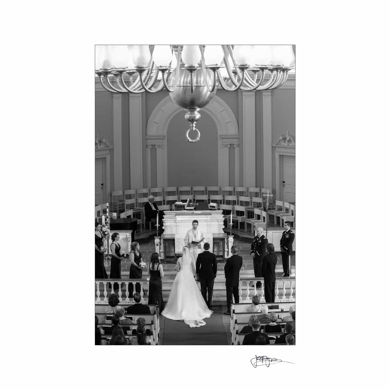 chapel hill wedding photographer - alexandra & john A black and white photo of a wedding ceremony inside a church. A couple stands at the altar, surrounded by the bridal party, with guests seated in pews. A large chandelier is visible above.
