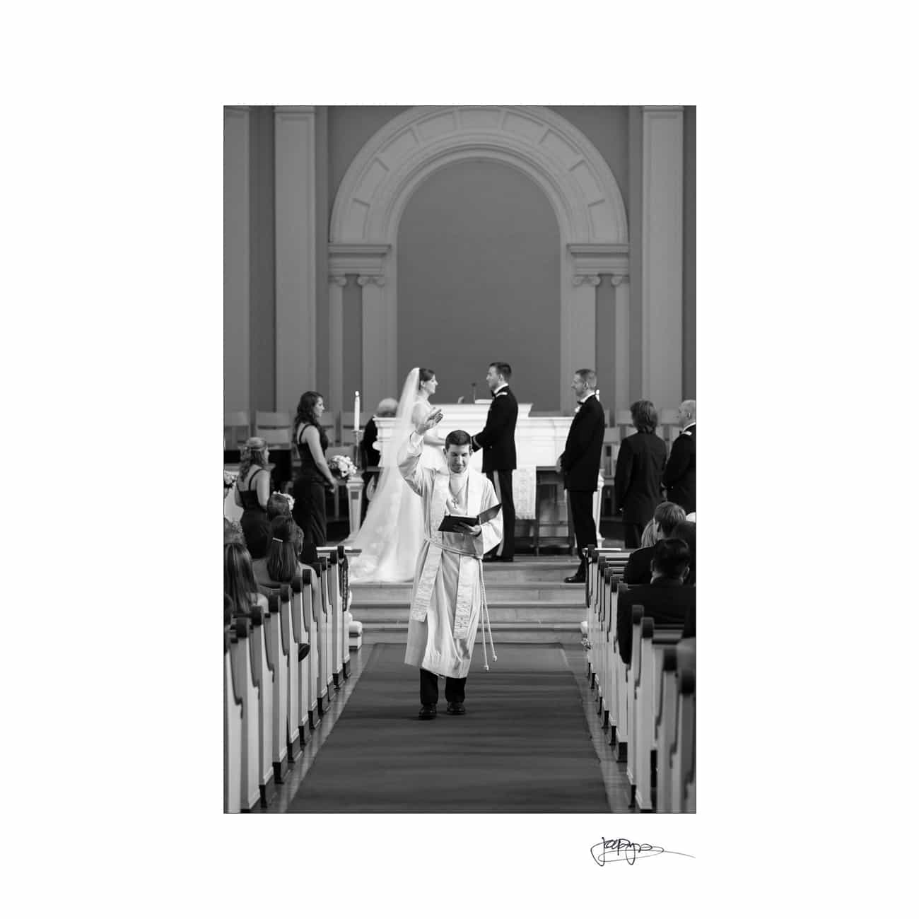 chapel hill wedding photographer - alexandra & john A black and white image of a wedding ceremony. The couple is in the background, embracing near the altar. A religious officiant in robes walks down the aisle, holding a book, with guests seated on both sides.