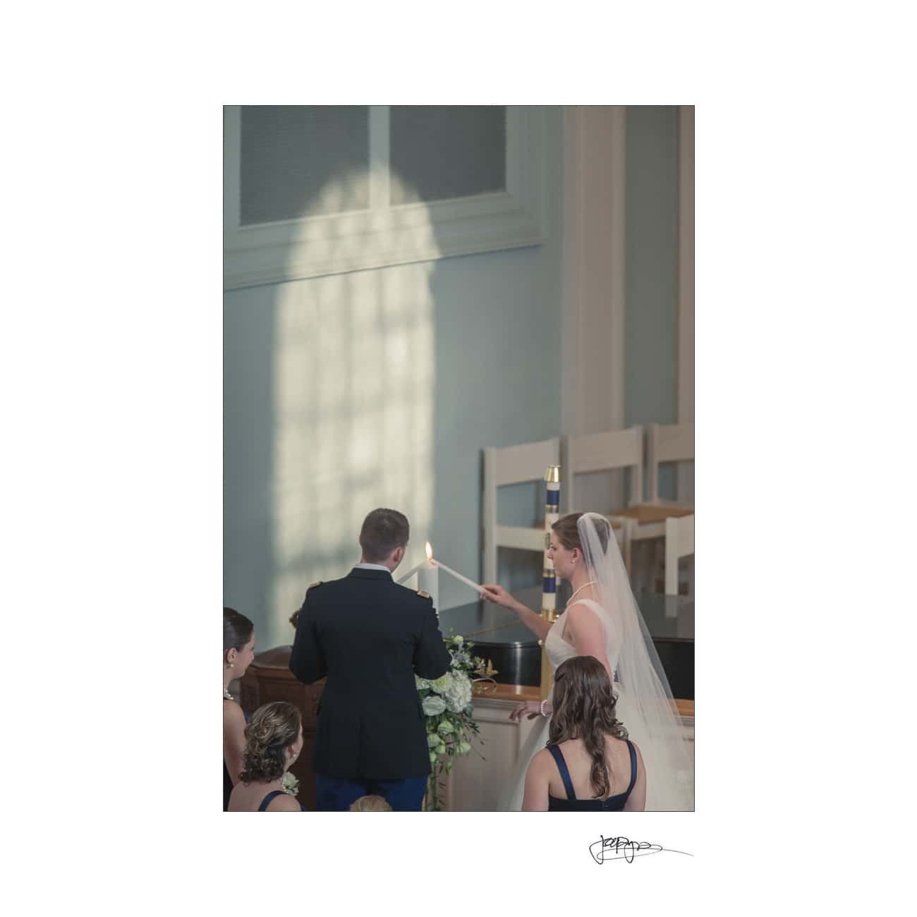 chapel hill wedding photographer - alexandra & john A bride in a white wedding dress and veil lights a candle with the groom in a suit. They stand together at an altar with bridesmaids in attendance, under soft natural light streaming through a window.