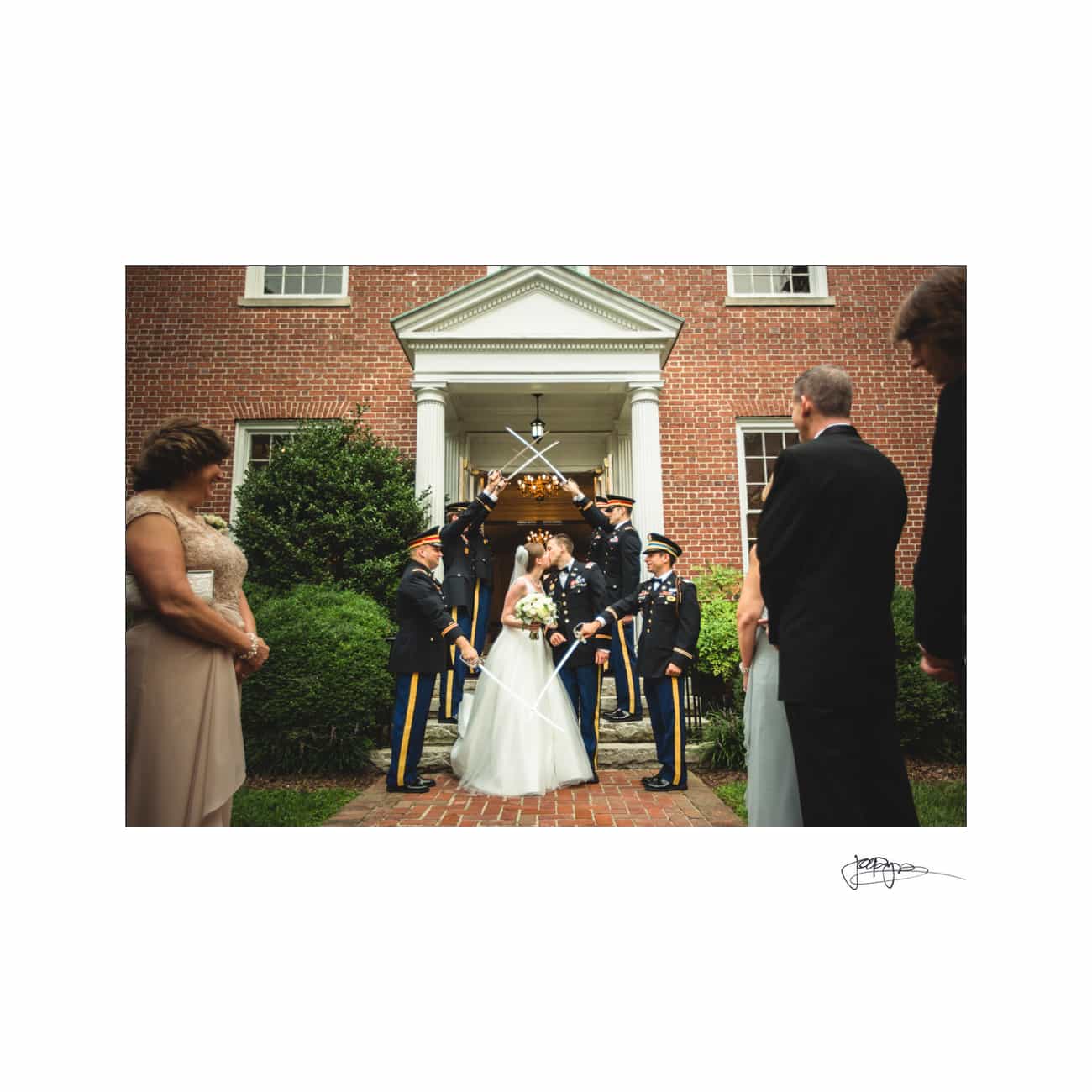 chapel hill wedding photographer - alexandra & john A bride and groom stand under an arch of swords held by uniformed officers at the entrance of a brick building. They are surrounded by guests in formal attire, with greenery and a formal doorway as the backdrop.