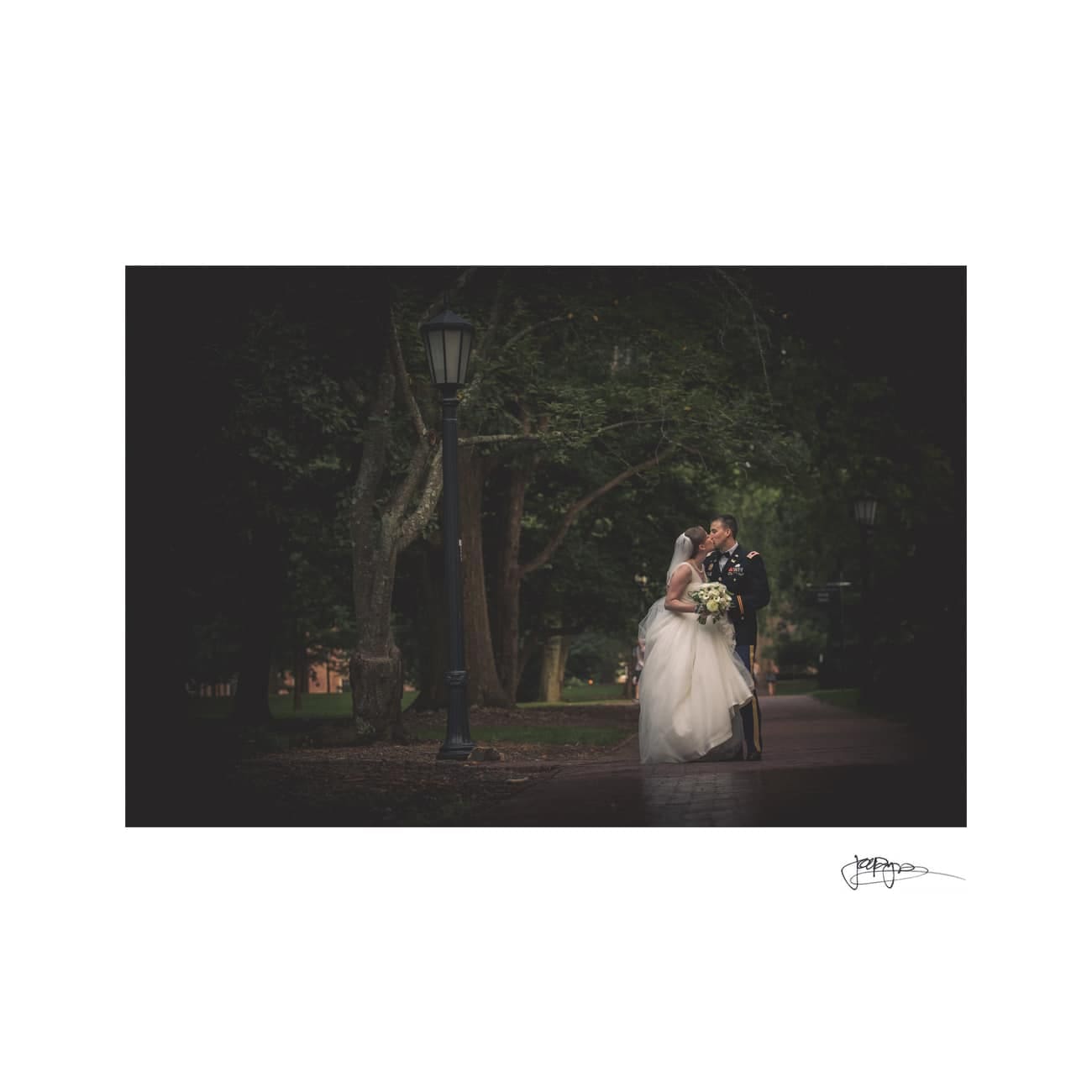 chapel hill wedding photographer - alexandra & john A bride and groom share a kiss under a lamppost on a tree-lined path. The bride holds a bouquet, and both are elegantly dressed. The scene is dimly lit, giving a romantic and intimate atmosphere.