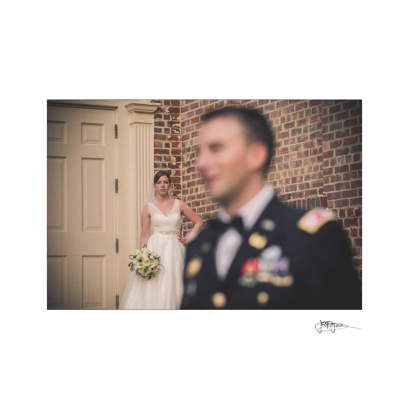 chapel hill wedding photographer - alexandra & john A bride in a white gown holding a bouquet stands against a brick wall near a door. In the foreground, a man in a military dress uniform is blurred. The focus is on the bride, creating a dramatic wedding scene.