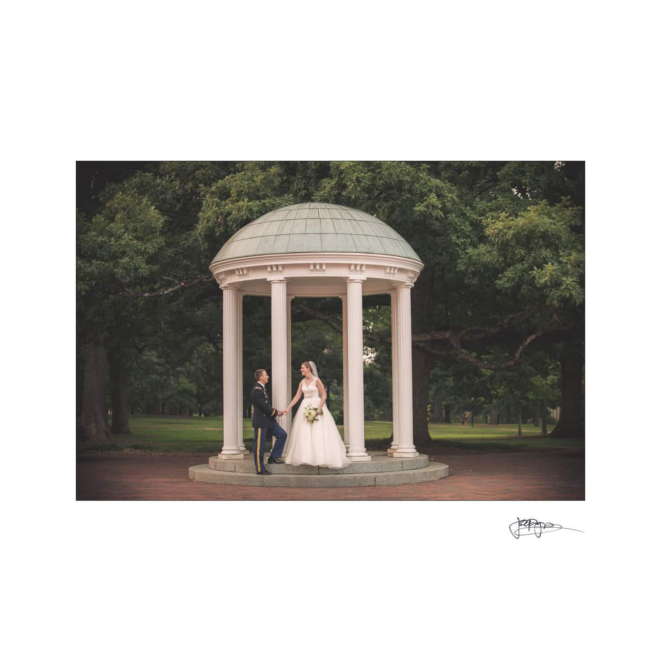 chapel hill wedding photographer - alexandra & john A bride and groom stand under a small, round, white pavilion with columns. The bride is in a white gown holding a bouquet, and the groom in a dark suit stands on the steps. Lush green trees are in the background.
