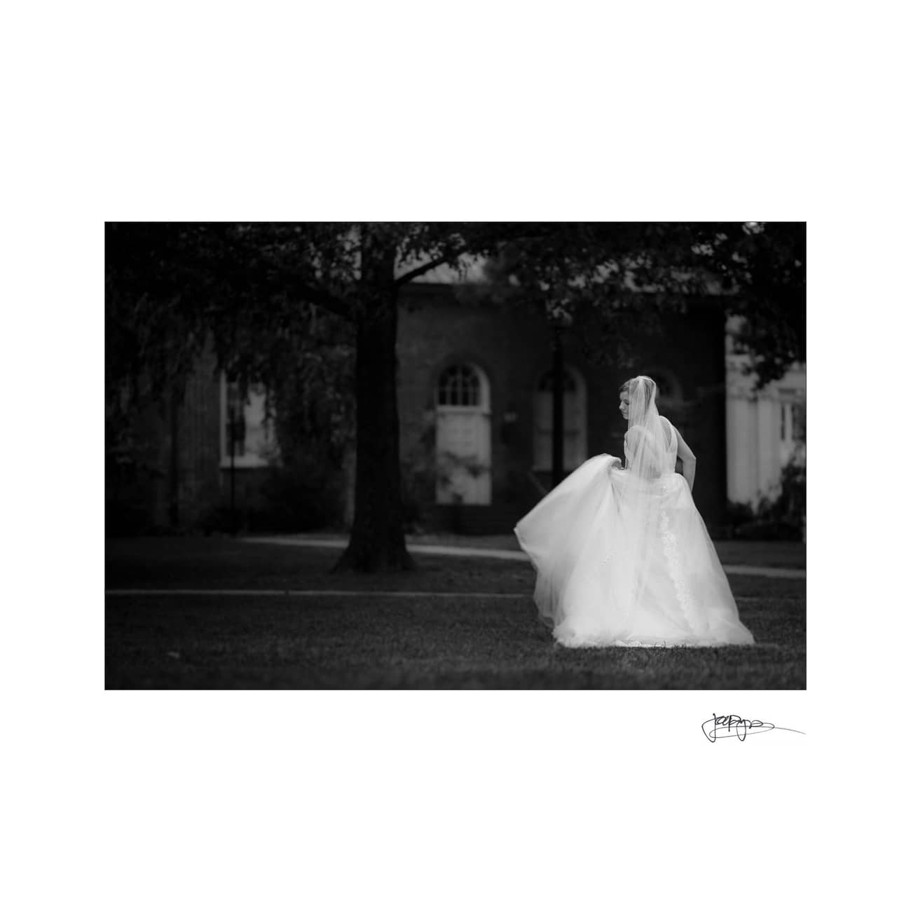 chapel hill wedding photographer - alexandra & john A black and white photo of a bride in a flowing gown walking on grass, with trees and a building in the background. The image captures a serene moment from a side angle.