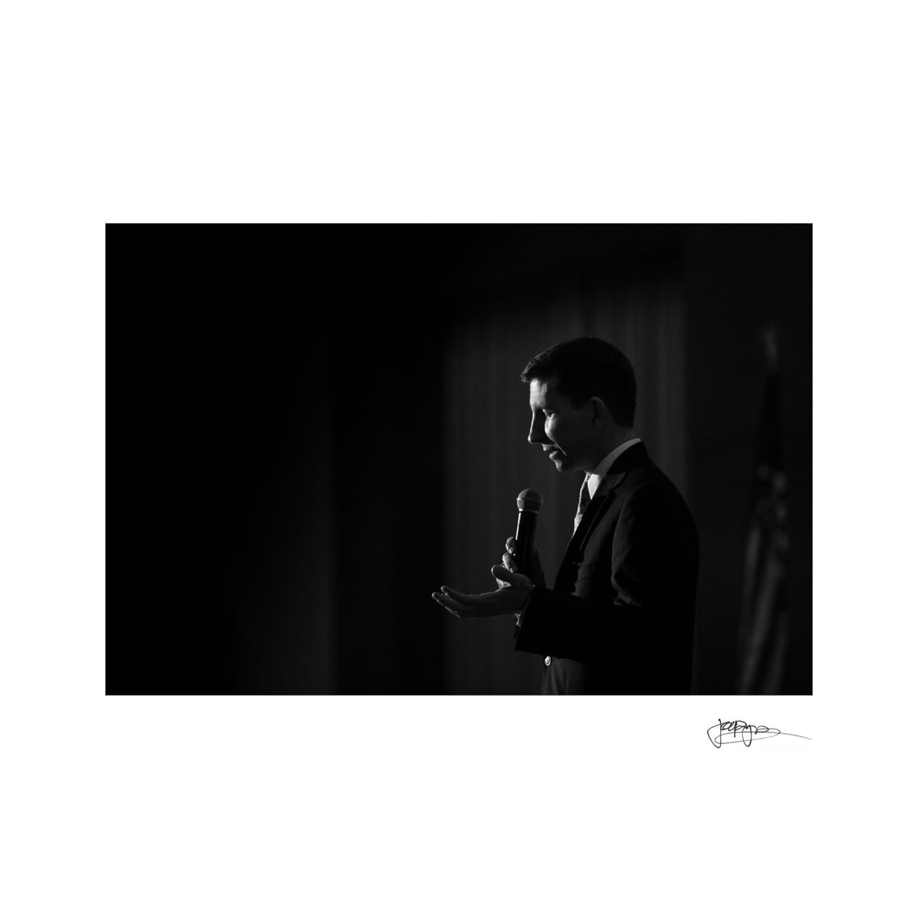 chapel hill wedding photographer - alexandra & john A person in a suit stands in profile, speaking into a microphone. The setting is dimly lit, creating a dramatic contrast between light and shadow. An indistinct flag is visible in the background.