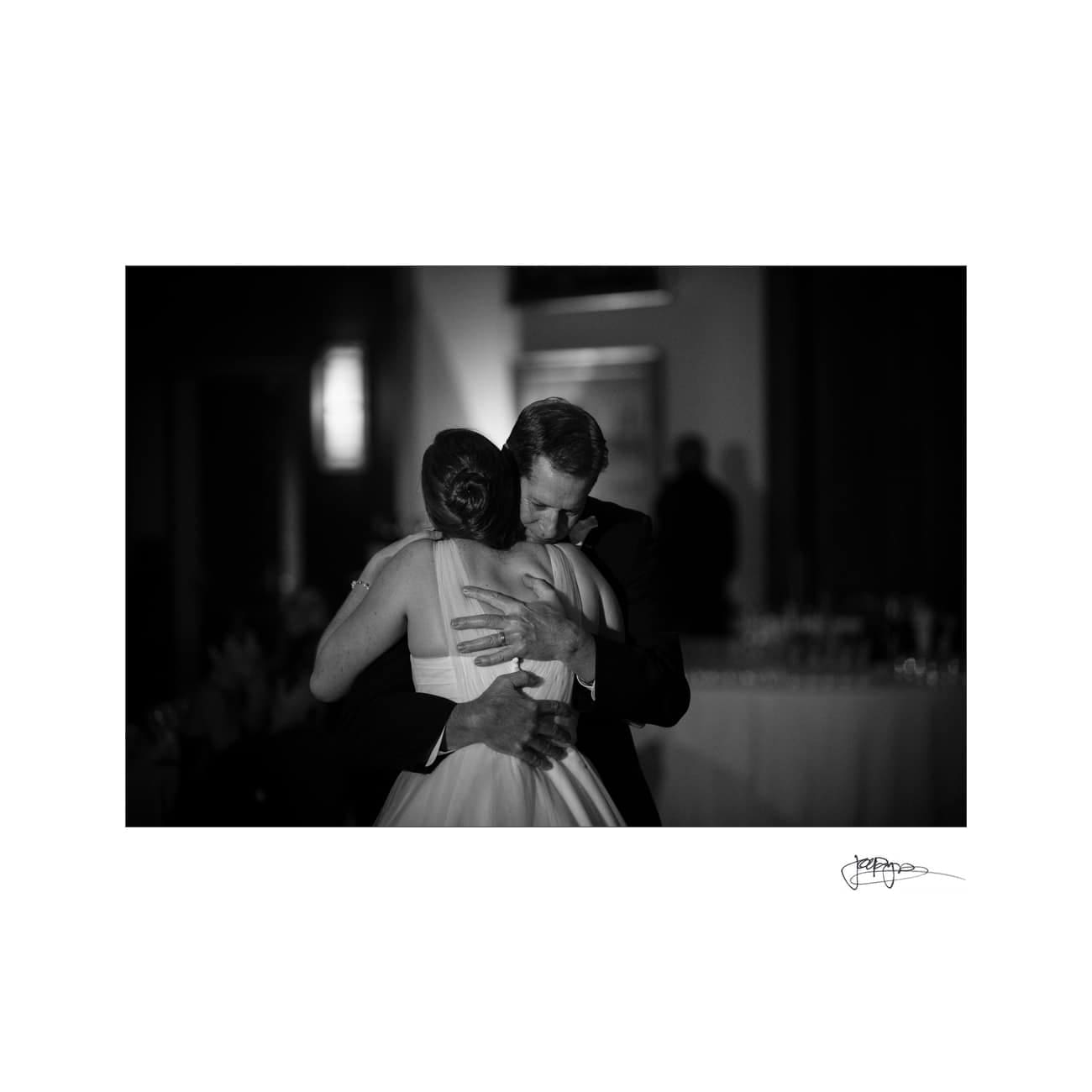 chapel hill wedding photographer - alexandra & john A black and white photo of two people embracing closely in a warm hug. The setting appears to be a formal event, with dim lighting in the background. The focus is on the emotional connection between the two individuals.