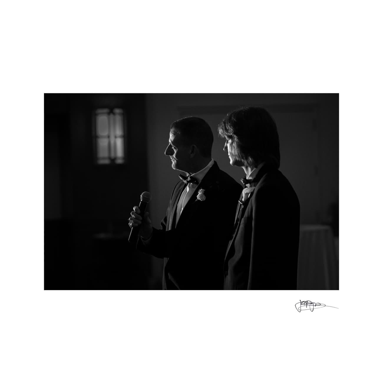 chapel hill wedding photographer - alexandra & john A black-and-white photo shows two people in formal attire. One is holding a microphone, possibly speaking or delivering a speech. The focus is on their side profiles, with dim lighting highlighting their faces.