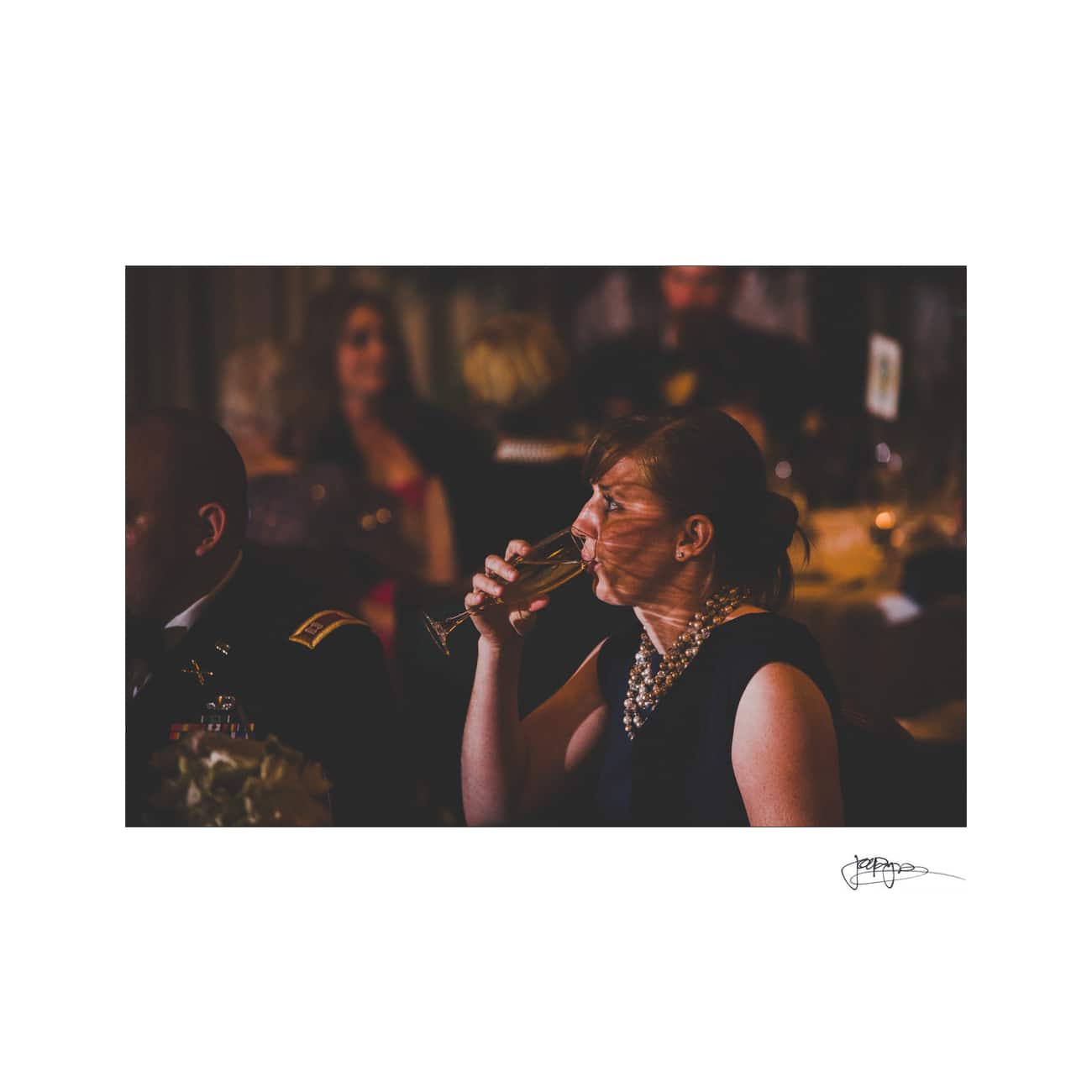 chapel hill wedding photographer - alexandra & john A woman in a dimly lit setting drinks from a champagne flute. She wears a pearl necklace and a dark dress. In the background, blurred figures sit at tables decorated with flowers, creating an elegant atmosphere.