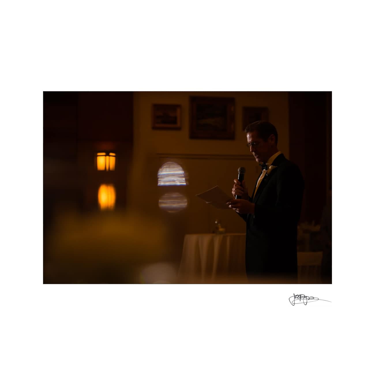 chapel hill wedding photographer - alexandra & john A person stands in dim lighting, holding a microphone and reading from a piece of paper. Elegant background with framed pictures and soft lighting adds a warm ambiance. Signs of a formal event visible.