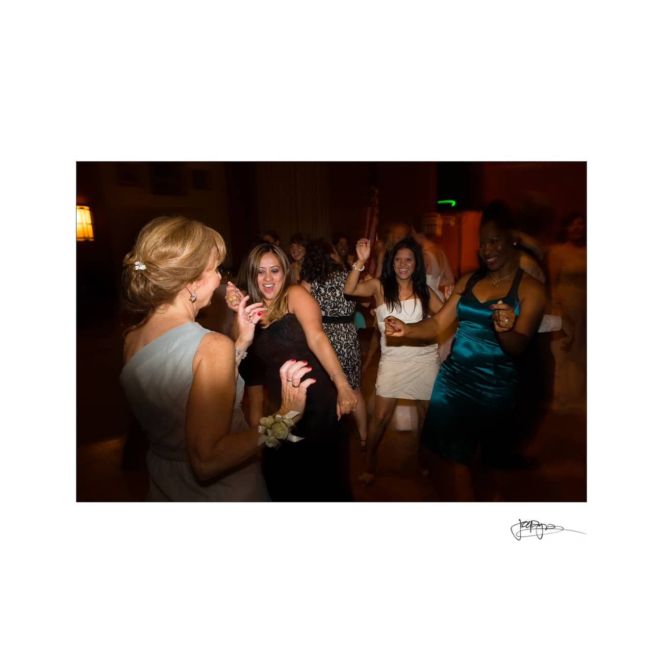 chapel hill wedding photographer - alexandra & john A group of women, dressed in elegant attire, joyfully dancing together in a dimly lit room. Some are smiling and raising their arms while wearing dresses in various colors, creating a lively and festive atmosphere.