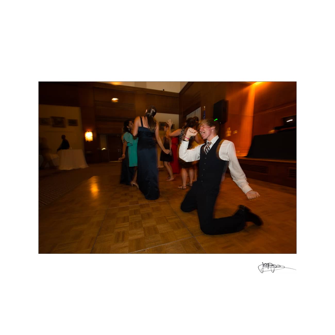 chapel hill wedding photographer - alexandra & john A group of people on a dance floor, with one man in a suit kneeling and striking a playful pose in the foreground. The background shows others also enjoying the dance, with dim lighting and a warm ambiance.
