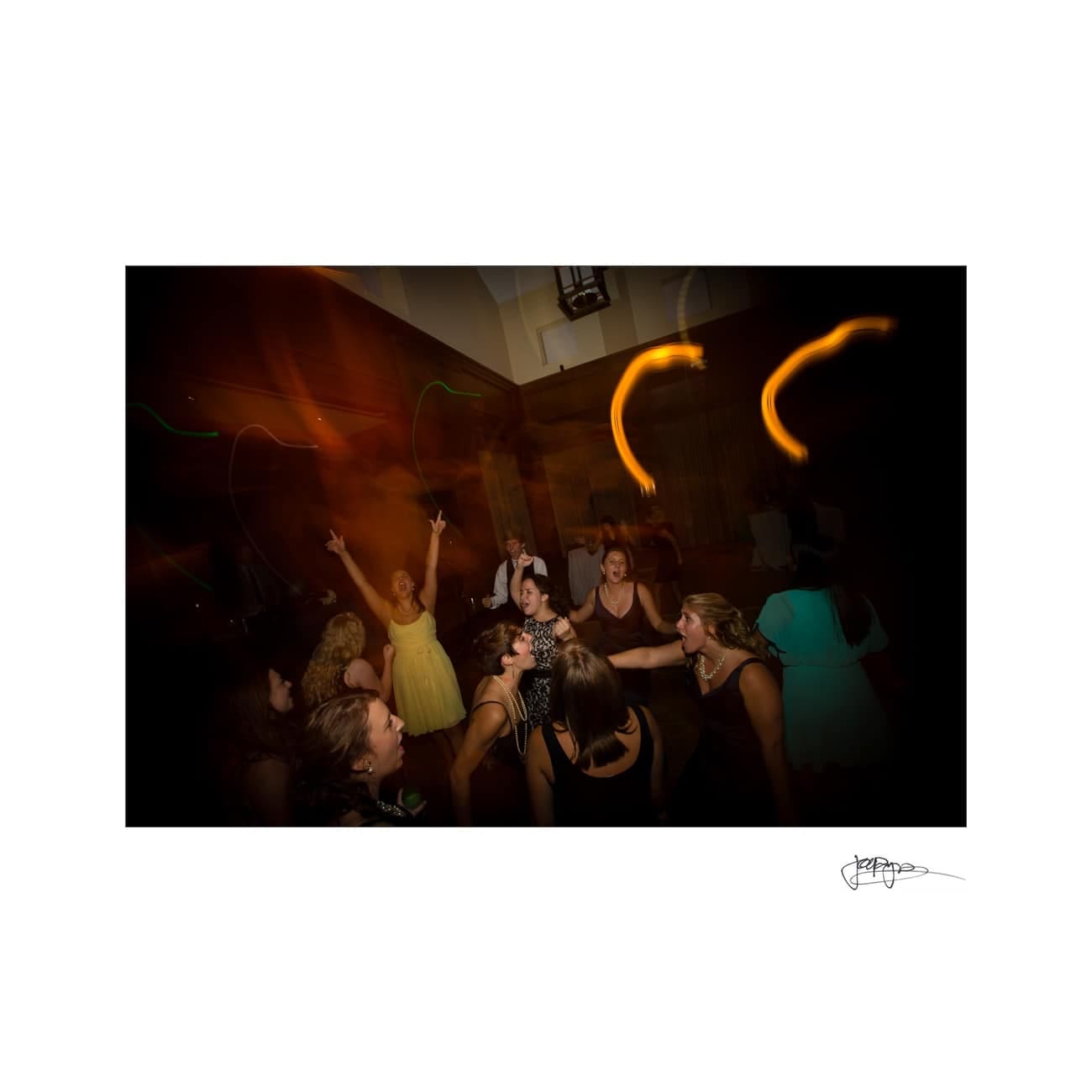 chapel hill wedding photographer - alexandra & john A group of people are dancing joyfully in a dimly lit room. The central figure in a yellow dress has their arms raised. Blurred lights create dynamic streaks above them, adding to the lively atmosphere.