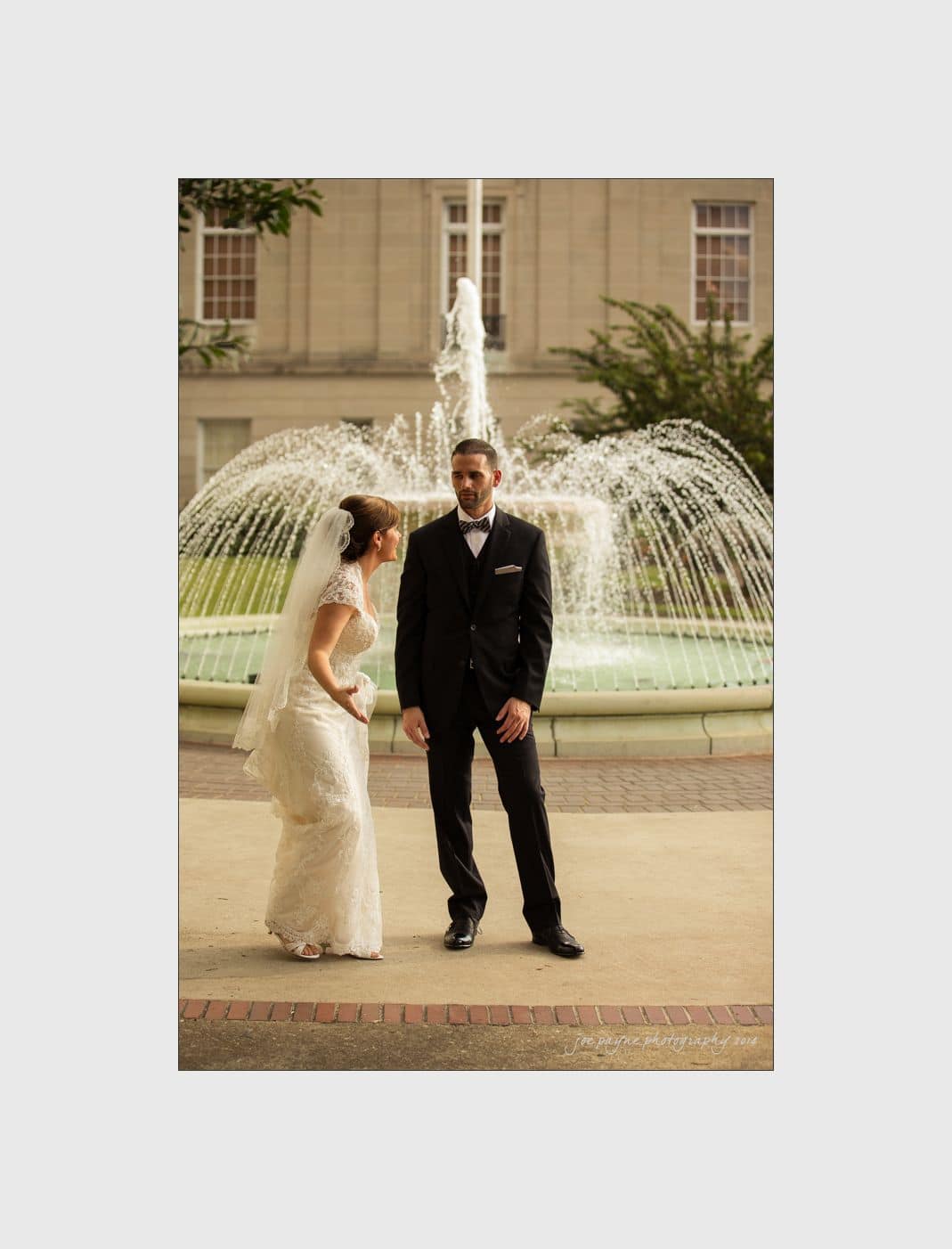 wilmington wedding photographer - nichole & eric A bride and groom stand in front of a large fountain. The bride is wearing a white dress with a veil, and the groom is in a black suit. Both appear to be smiling and are posed casually against the backdrop of a building.