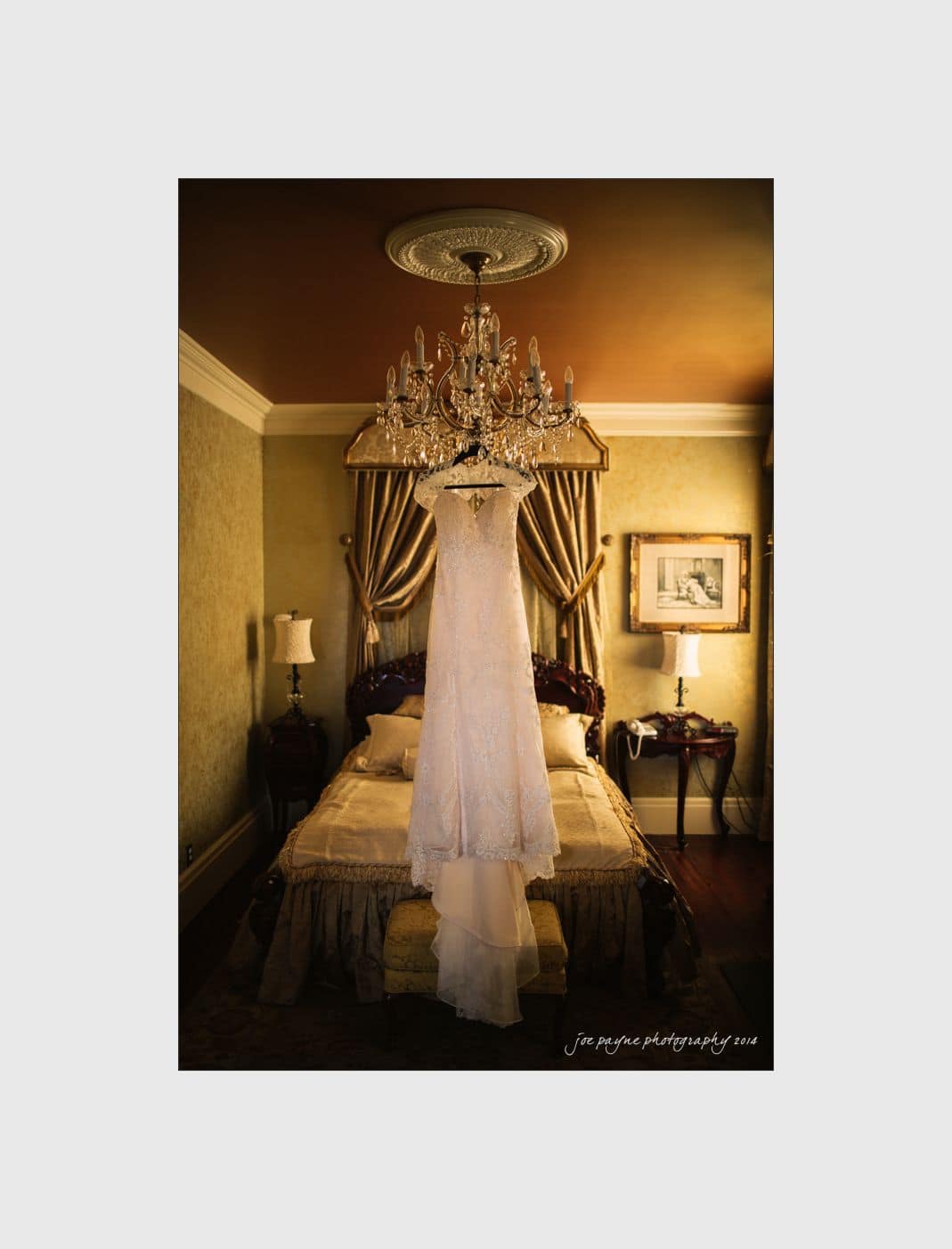 wilmington wedding photographer - nichole & eric A white wedding dress hangs from a chandelier above a luxurious, vintage-style bedroom. The room features elegant drapery, a framed picture on the wall, and soft lighting that highlights the intricate details of the dress.