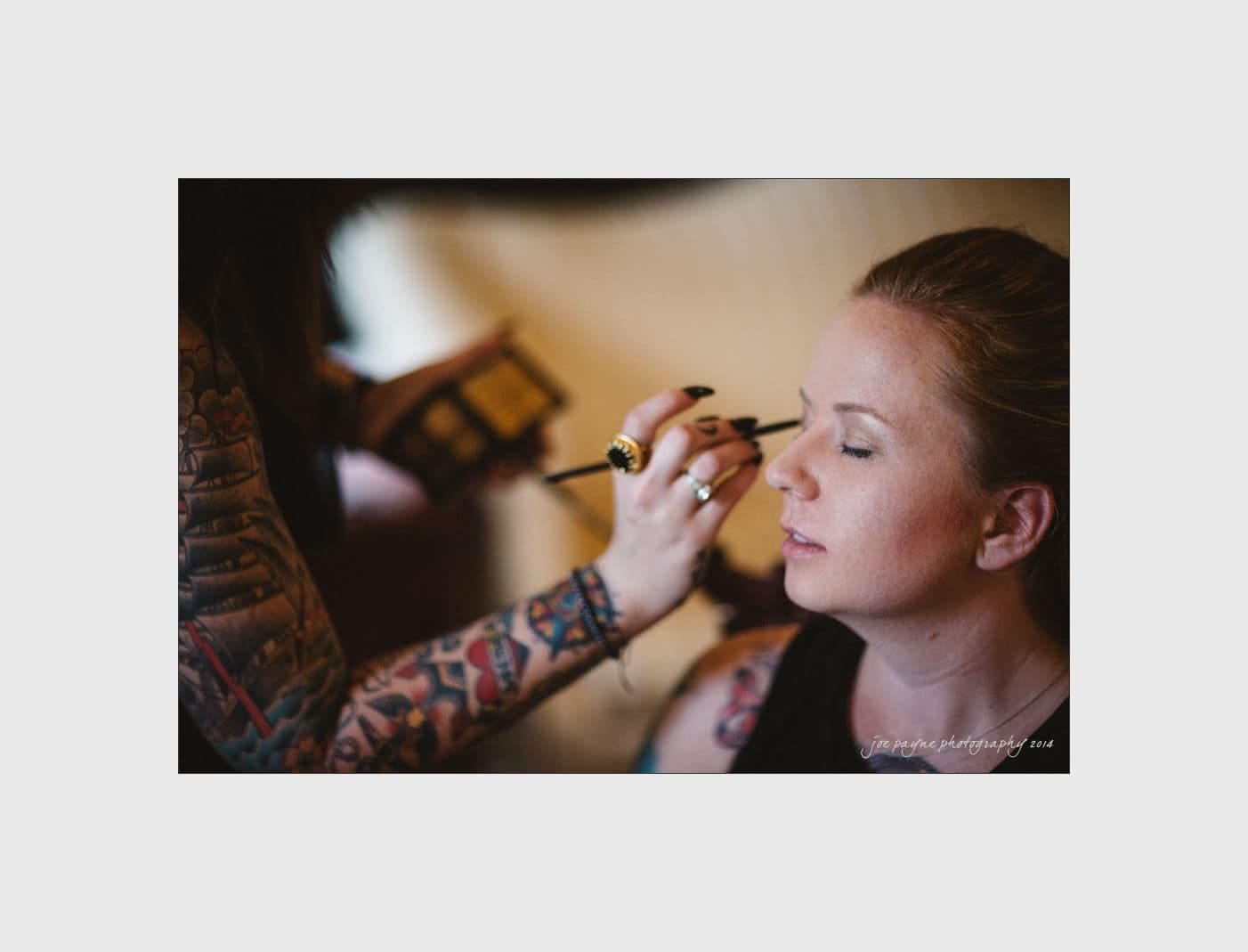 wilmington wedding photographer - nichole & eric A person with tattoos on their arms is applying eye makeup with a brush to a seated woman who has her eyes closed. The background is softly blurred, creating a focused and intimate moment.