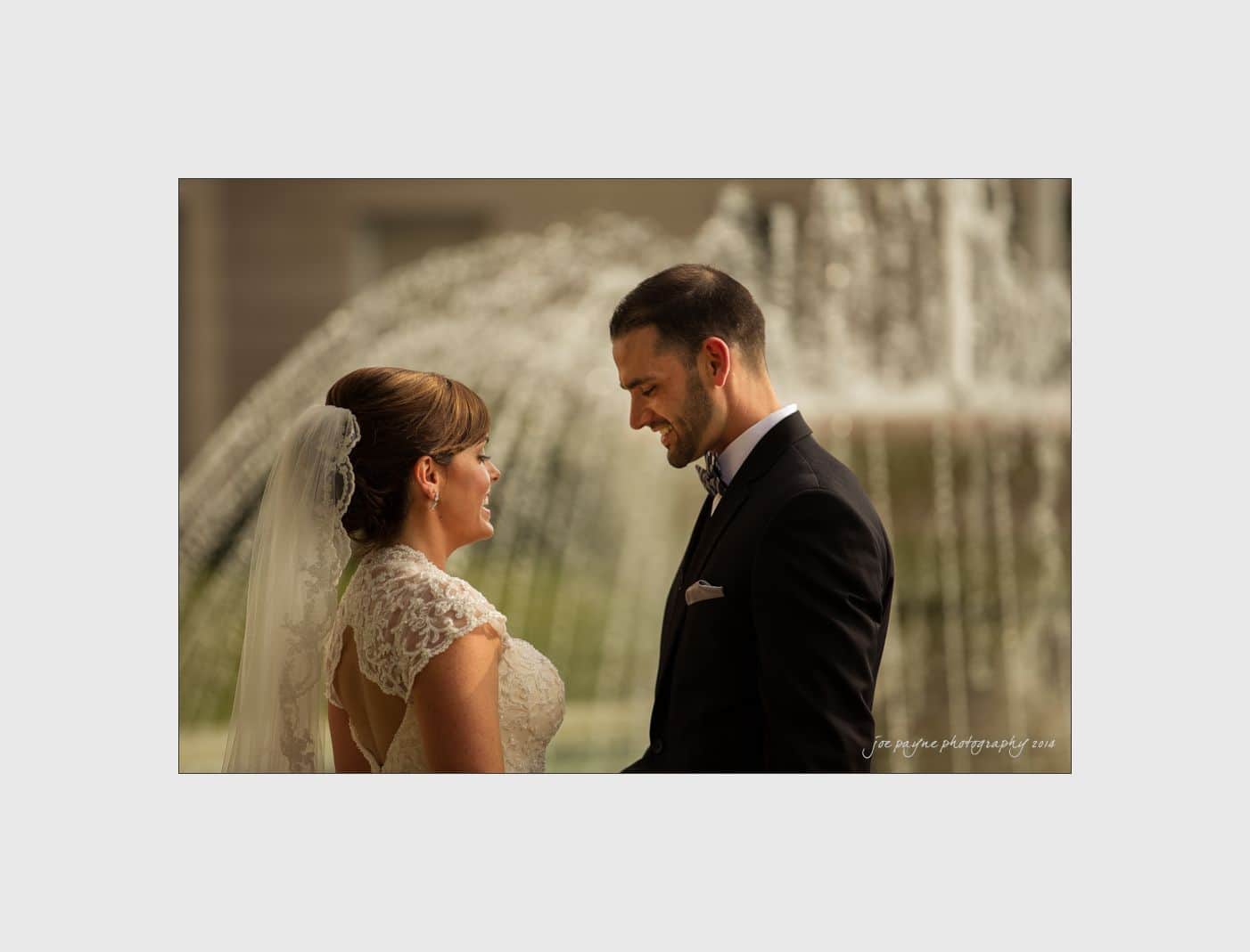 wilmington wedding photographer - nichole & eric A bride and groom stand facing each other, smiling in front of a large, flowing fountain. The bride wears a lace gown and veil, while the groom is in a dark suit with a bow tie.
